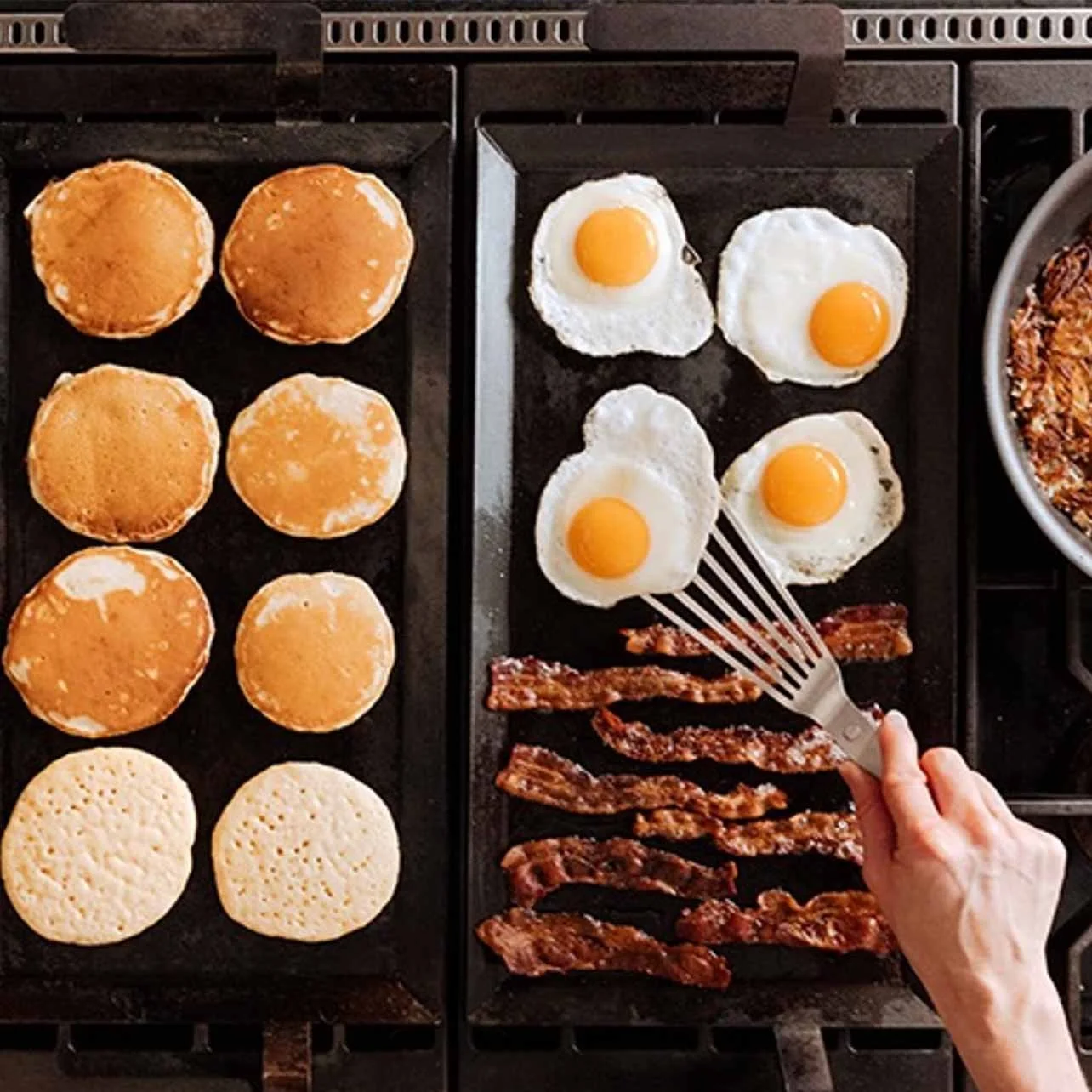Cooking breakfast with pancakes, sunny side up eggs, and bacon on stovetop griddles.
