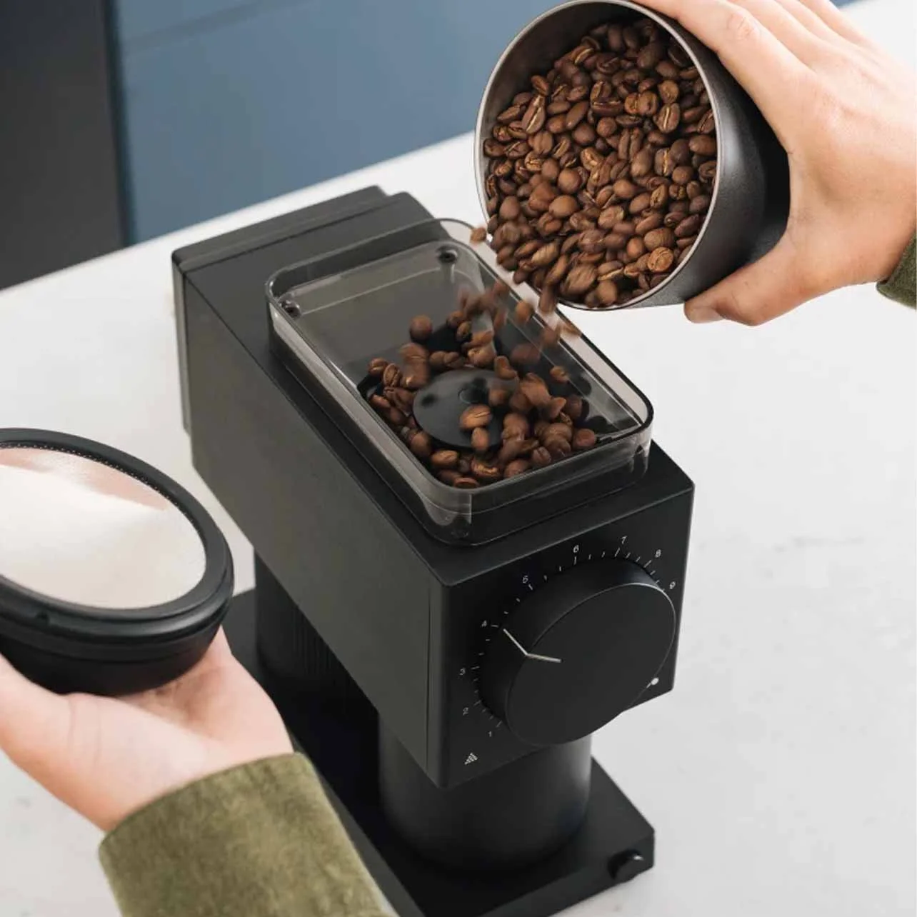 Person pouring roasted coffee beans into a coffee grinder while holding a container of coffee grounds.