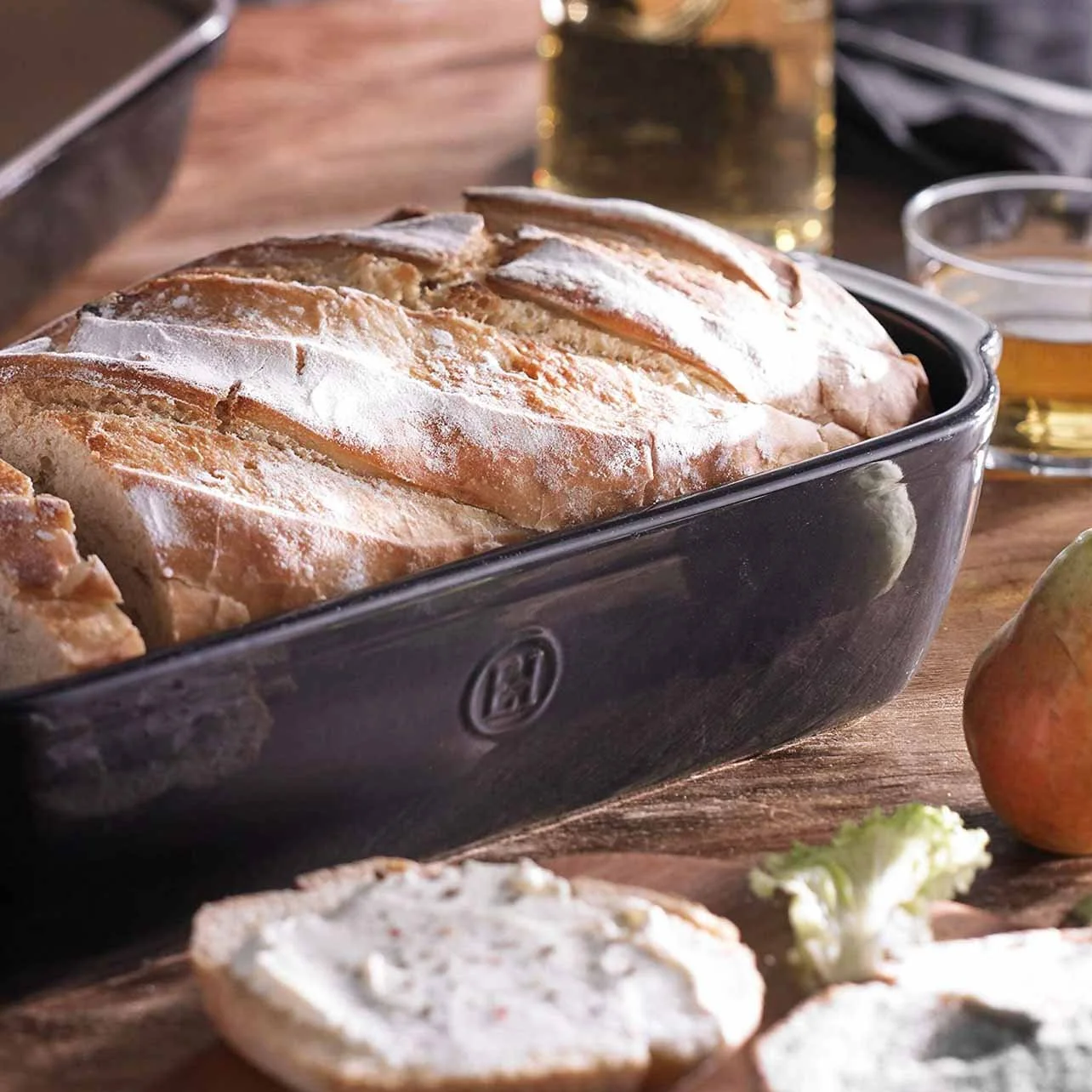 A loaf of bread in a black baking dish on a wooden table, with a glass of beer and a bottle of oil in the background, and some sliced bread and a pear in the foreground.