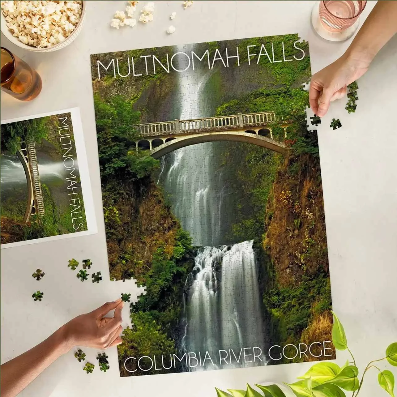 A person is assembling a jigsaw puzzle of the Columbia River Gorge waterfall scene. Several loose puzzle pieces are on a white surface, with a bowl of popcorn and glasses of drinks nearby.