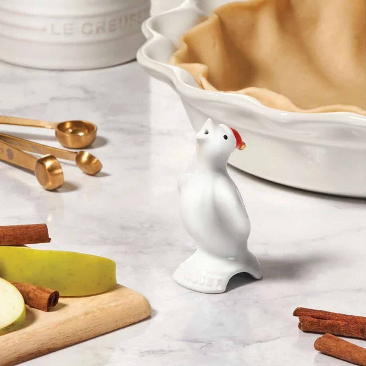 Close-up of a white ceramic polar bear figurine with a Santa hat, surrounded by cinnamon sticks, yellow apple slices, gold measuring spoons, and an apple pie with a golden crust on a white marble countertop.