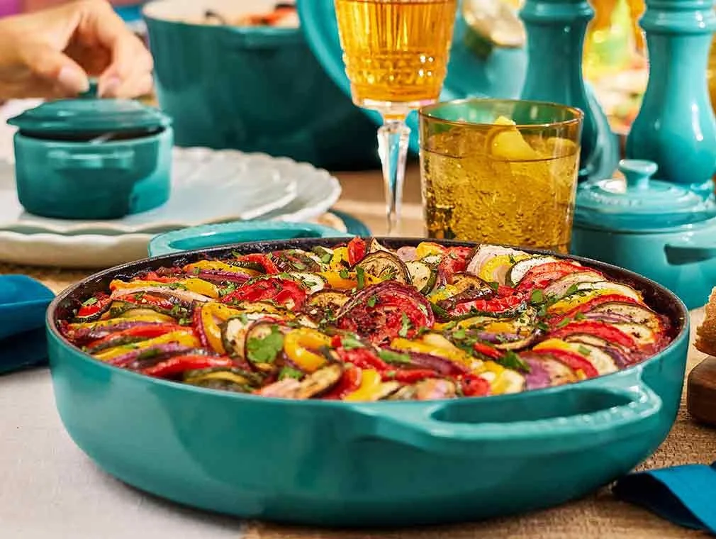 Colorful vegetable gratin in a turquoise dish on a dining table with drinks and tableware.