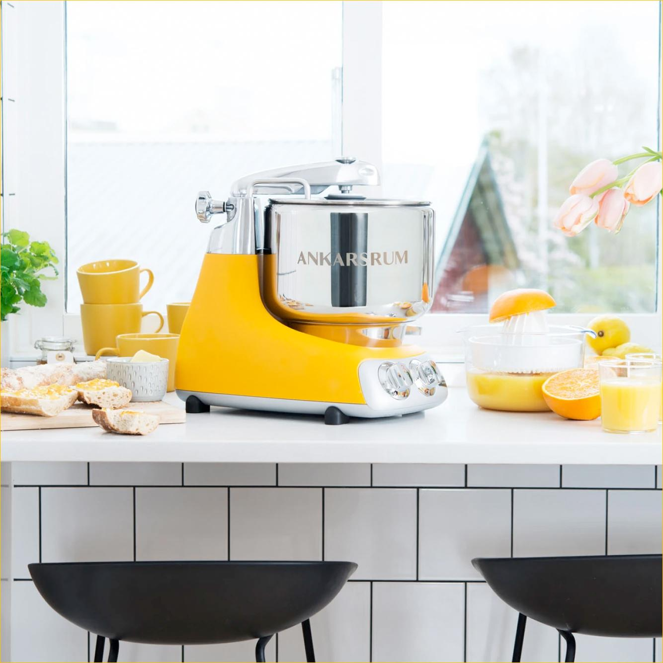 A retro-style yellow and stainless steel blender on a white kitchen counter with cups, bread, orange slices, and a bowl of orange juice, near a large window with pink tulips.