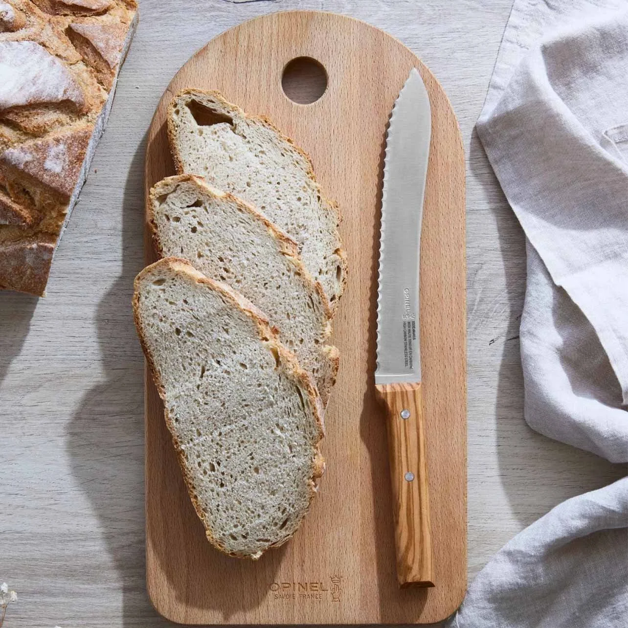 Three slices of rustic bread on a wooden cutting board with a serrated bread knife, a white linen cloth to the right, and part of a loaf of bread in the top left corner.