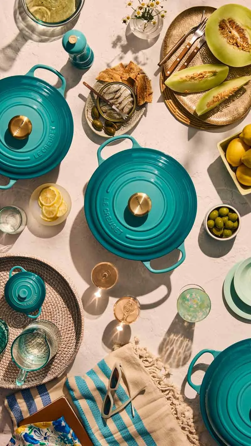 A colorful outdoor table set for a meal featuring turquoise Le Creuset cookware, sliced melons, lemons, olives, and a small vase with flowers, with sunlit shadows cast across the table.