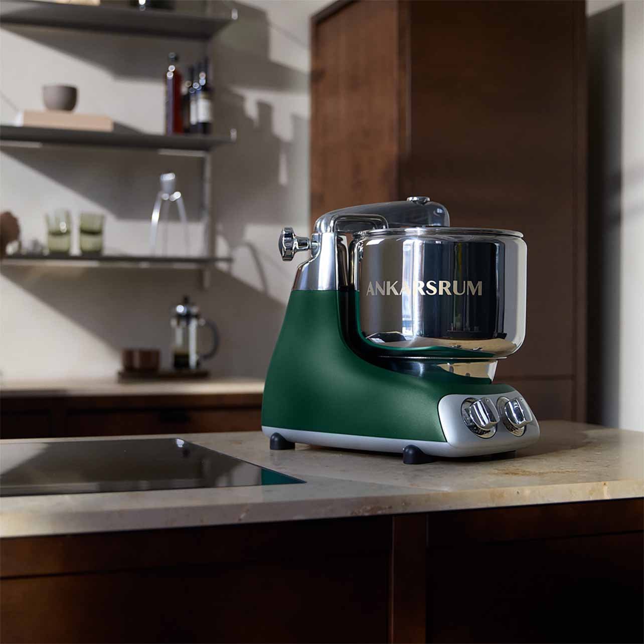 A vintage green and silver Ankarsrum stand mixer on a kitchen countertop with wooden cabinets and open shelves in the background.