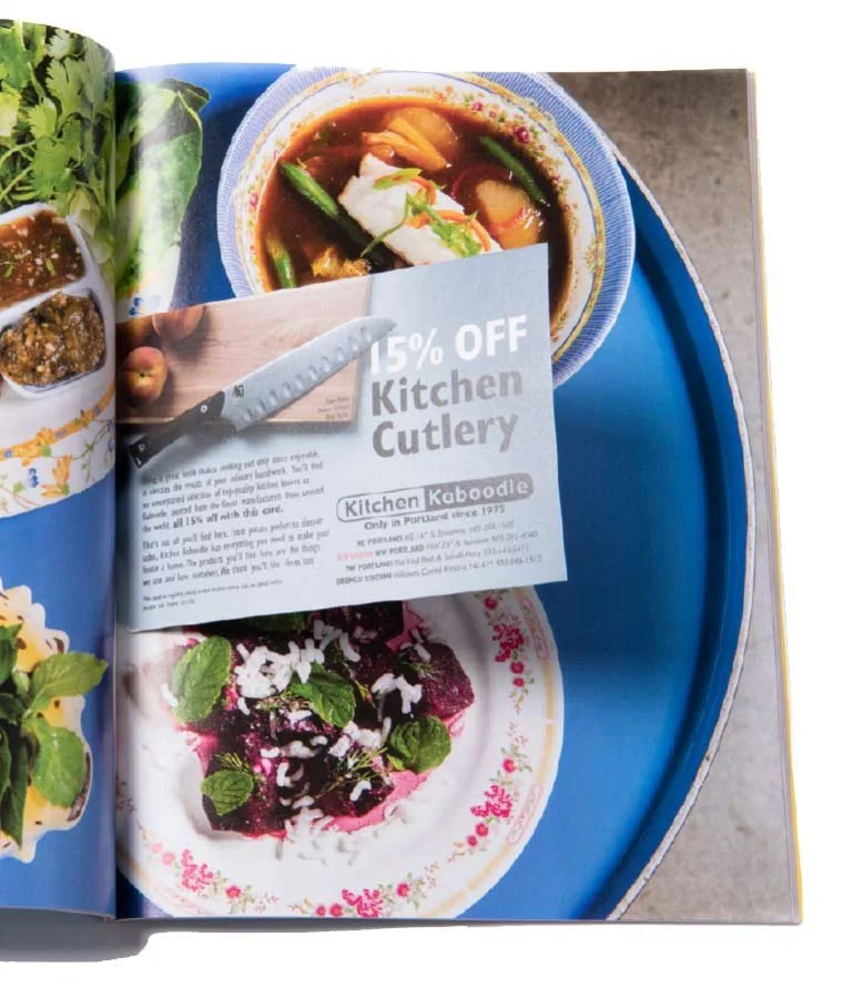 A magazine page showing a bowl of soup with vegetables and a plate of purple berries topped with mint leaves, and a promotional flyer for kitchen cutlery discounts.