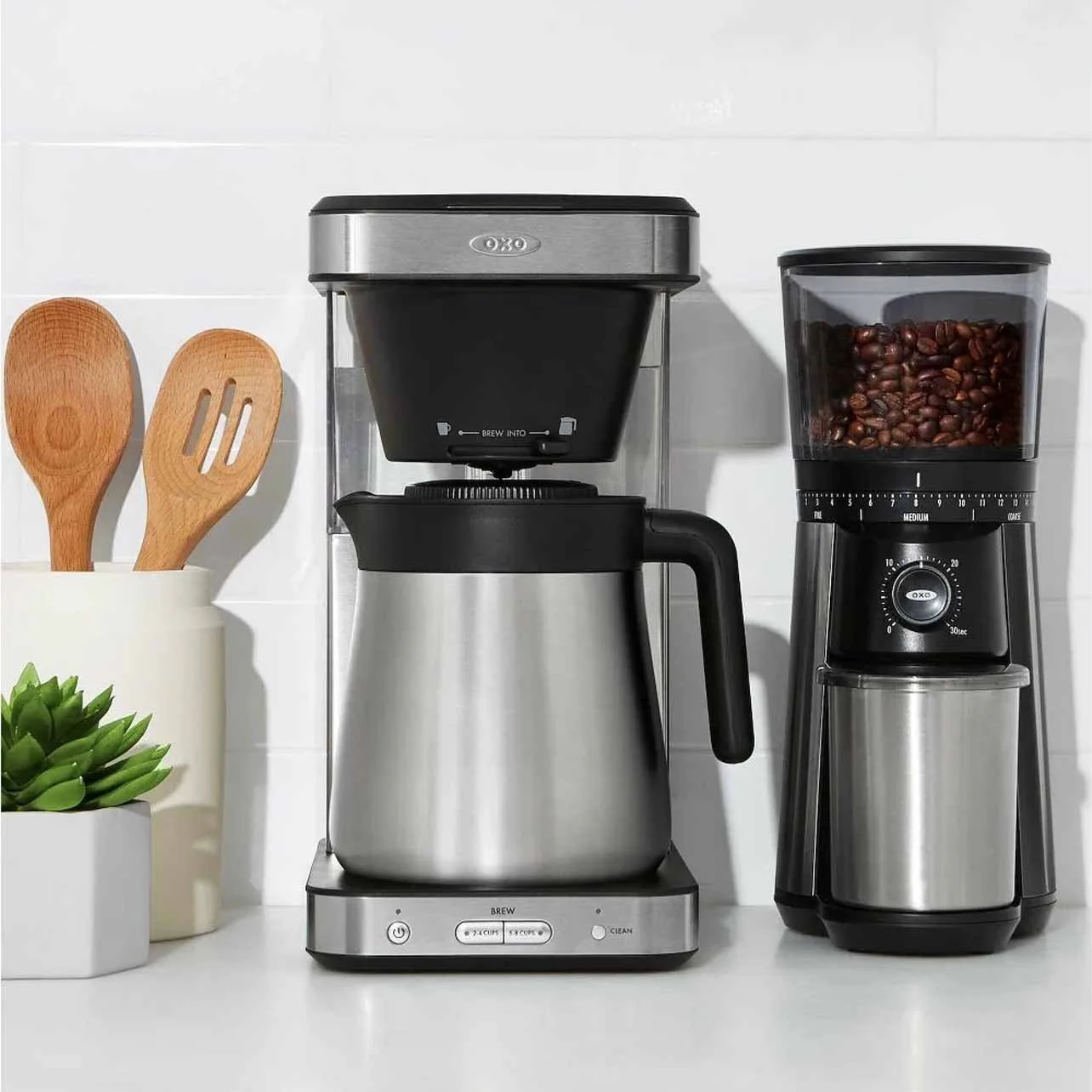 Countertop with coffee maker, coffee grinder filled with coffee beans, kettle, wooden utensils in a white container, and a small potted green plant.