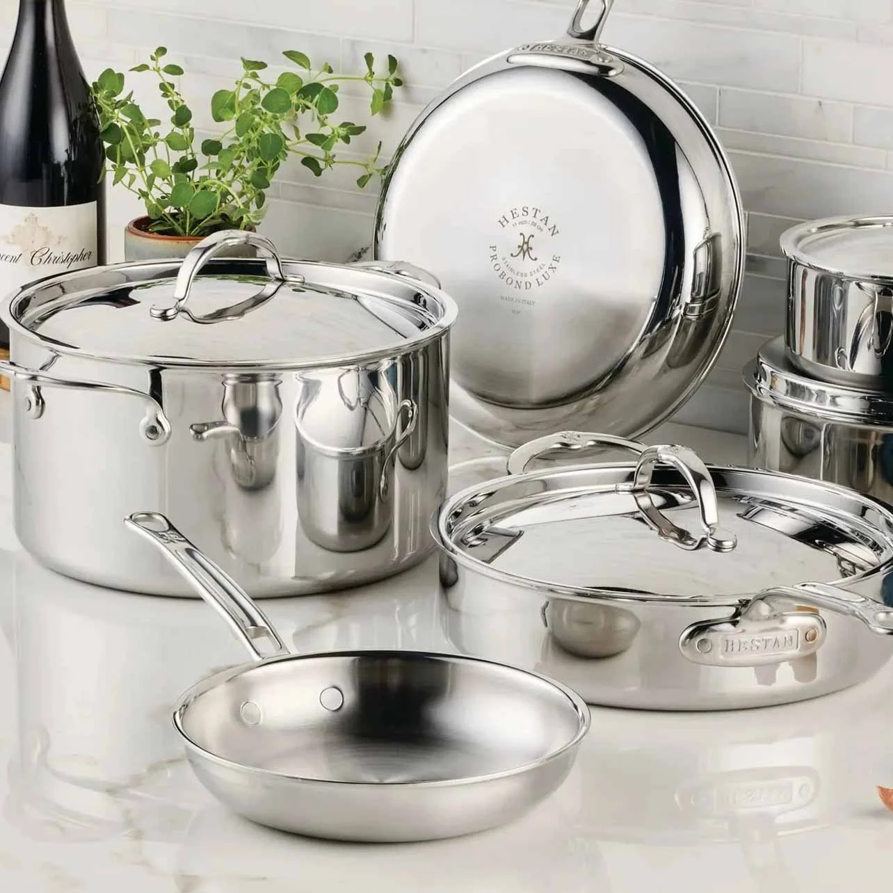 Set of shiny stainless steel pots and pans with lids on a white countertop. A potted plant and a bottle of wine are in the background.