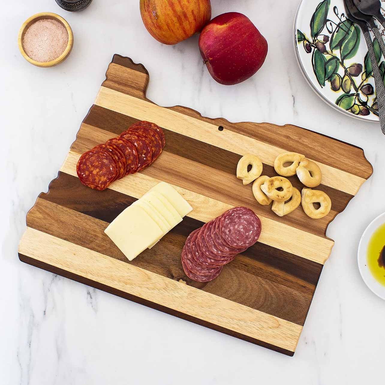 A wooden serving board shaped like the state of Ohio, with slices of salami, cheese, and pepperoni on it. There are two apples, a small bowl of pink seasoning, and a floral plate with utensils partially visible nearby.