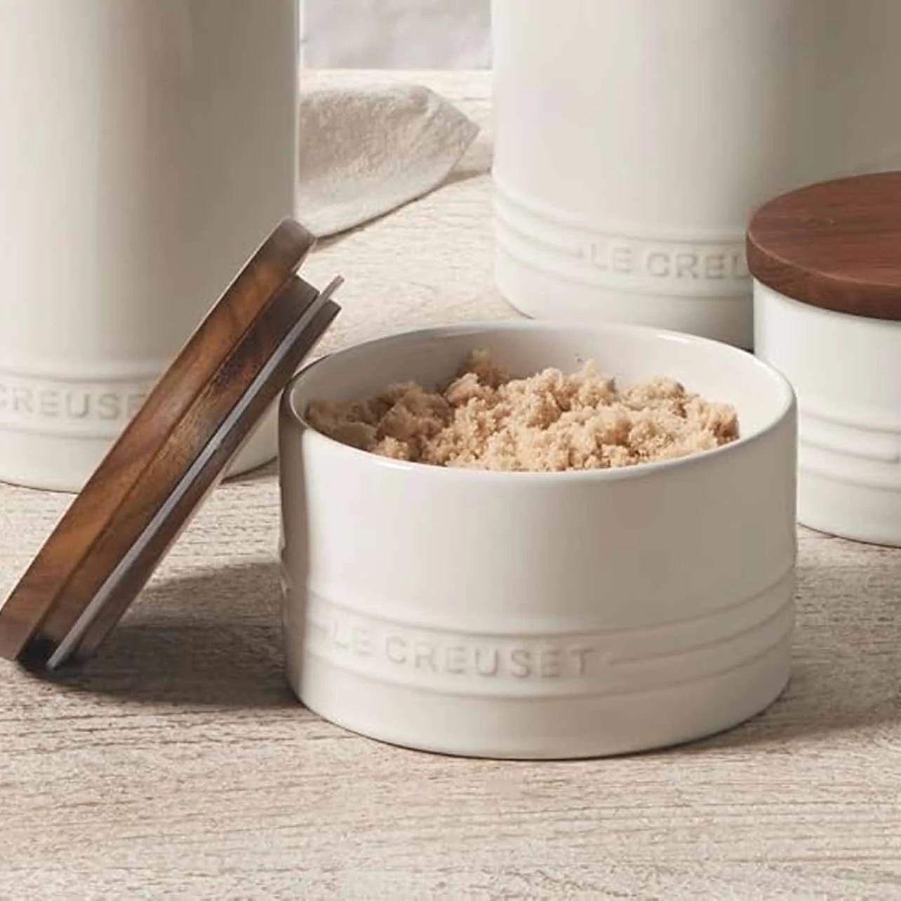 A round white container with the Le Creuset brand embossed on it, filled with brown sugar on a wooden surface, with other similar containers in the background.