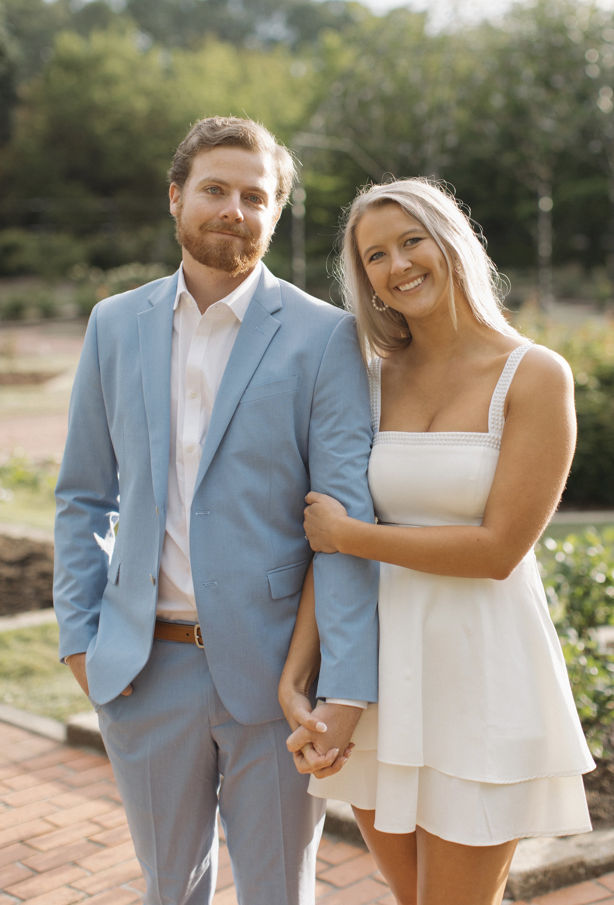 A smiling couple standing outdoors, holding hands. The man wears a light blue suit, and the woman wears a white dress, both in a park-like setting with trees and greenery.