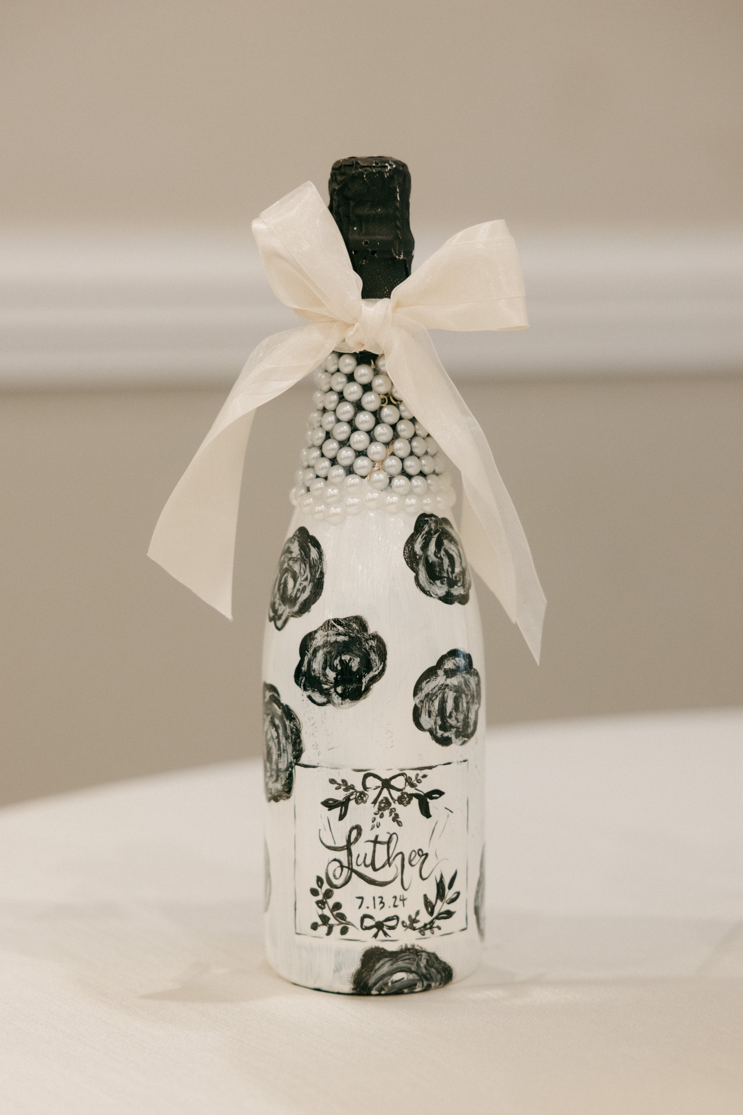 Decorated champagne bottle with black and white floral patterns, pearl embellishments, a cream ribbon bow, and a personalized label with the name Luther and date 7.13.24.