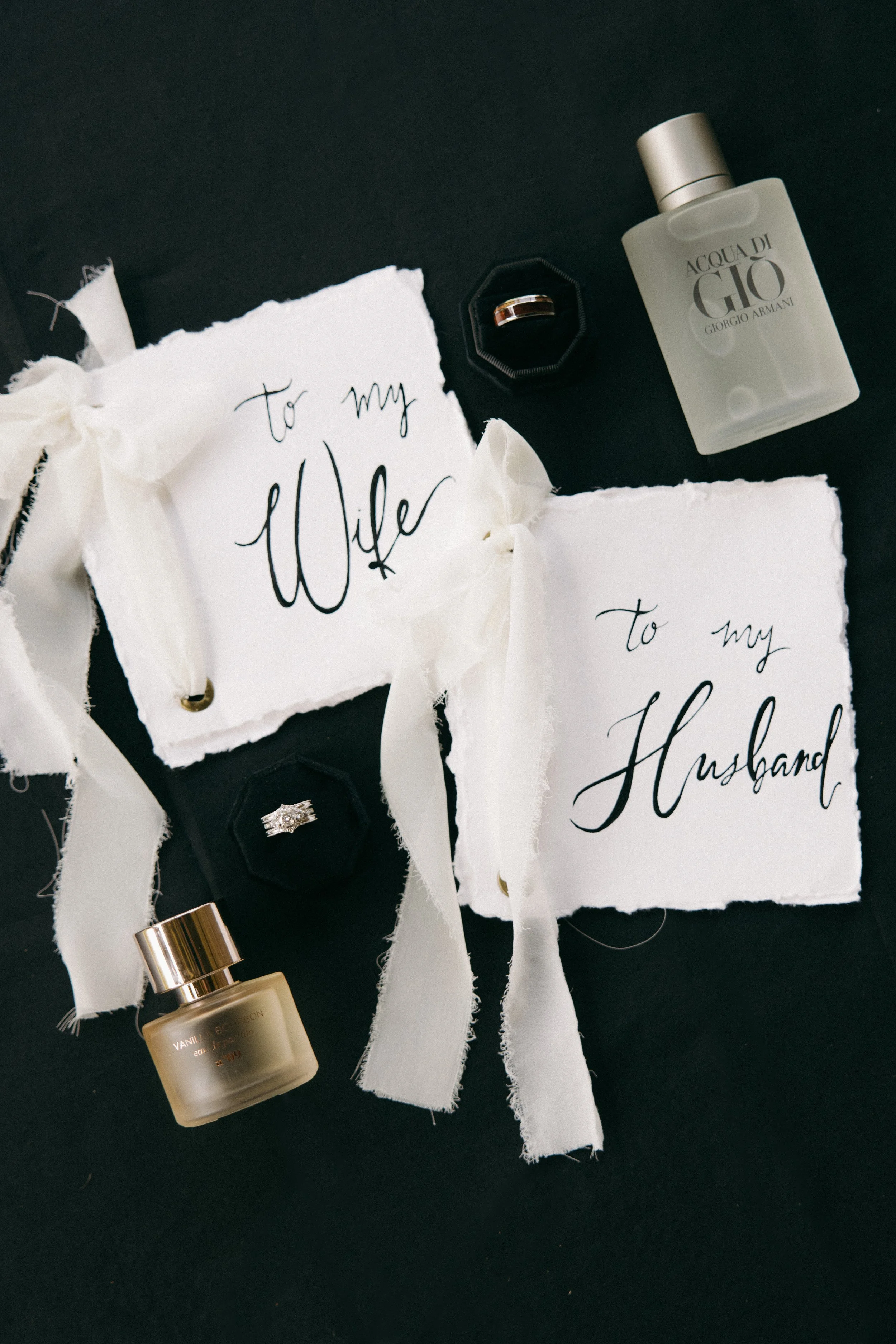 Wedding gifts including Monogrammed cards for wife and husband, rings, and perfume bottles on a black background.