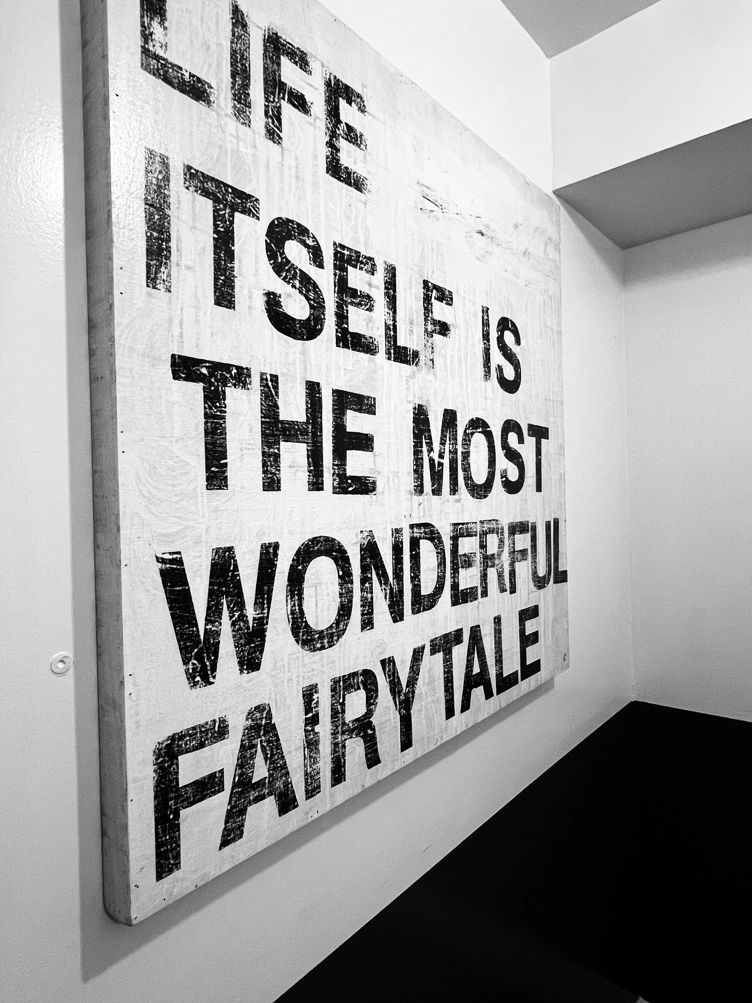 Black and white sign with the quote 'Life itself is the most wonderful fairy tale' written on a wooden background hanging on a white wall.