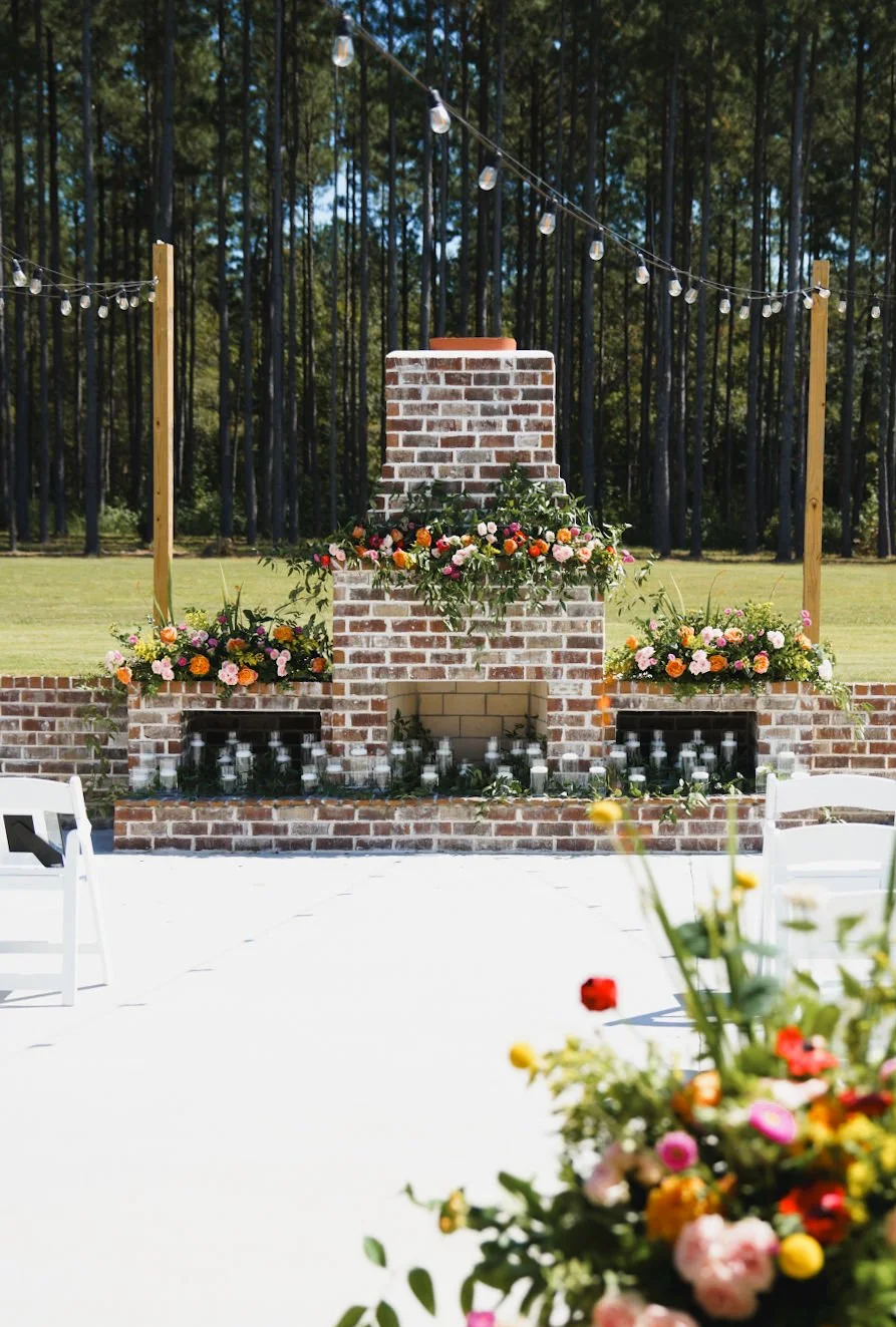 Outdoor wedding ceremony setup featuring a brick fireplace adorned with colorful flowers, string lights overhead, and greenery in a wooded area.