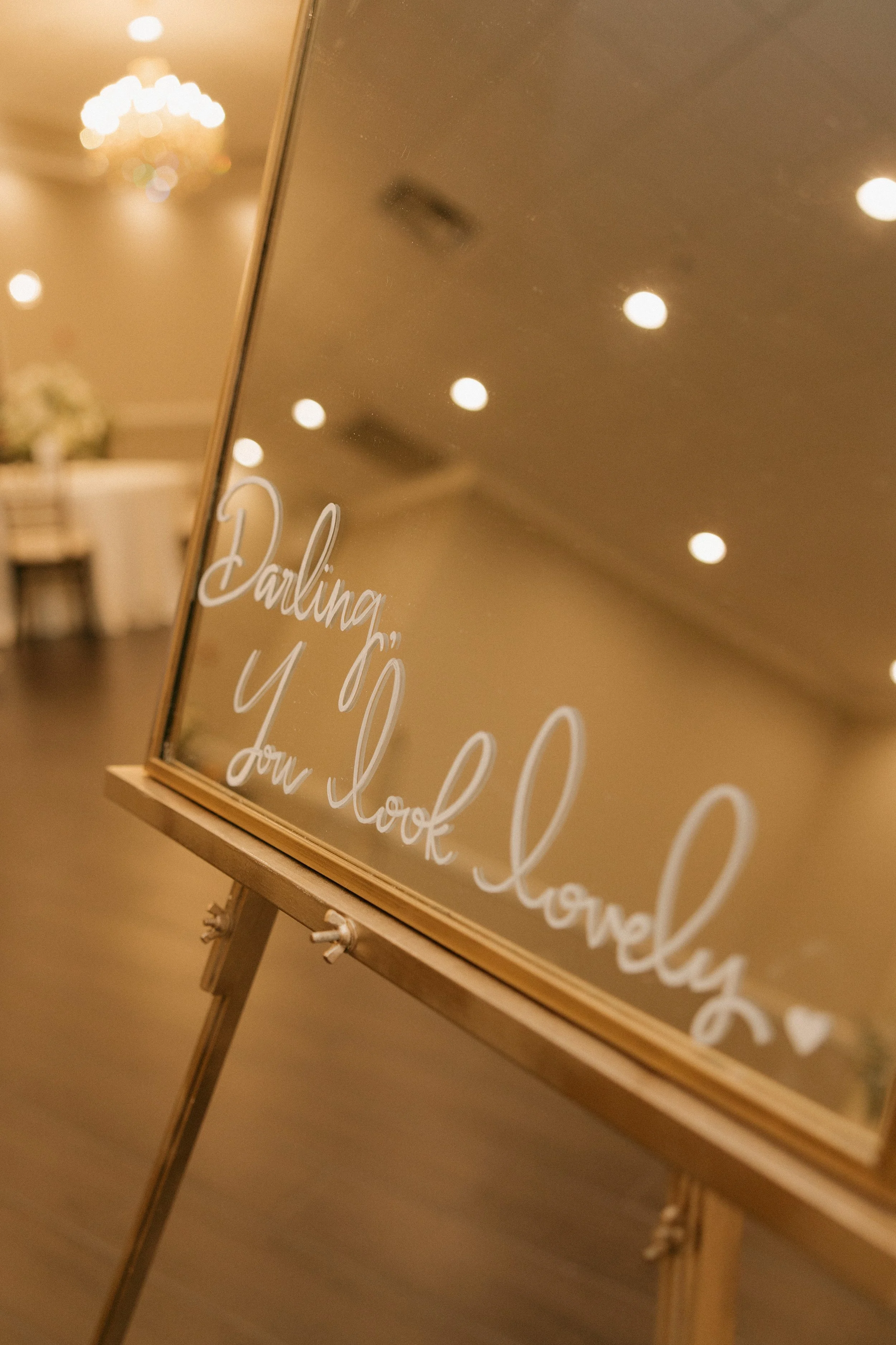 A mirror with a wooden frame on an easel displaying the message 'Daling, y your look lovely' written in white cursive text, with a blurry background of a decorated indoor space with hanging lights and tables.