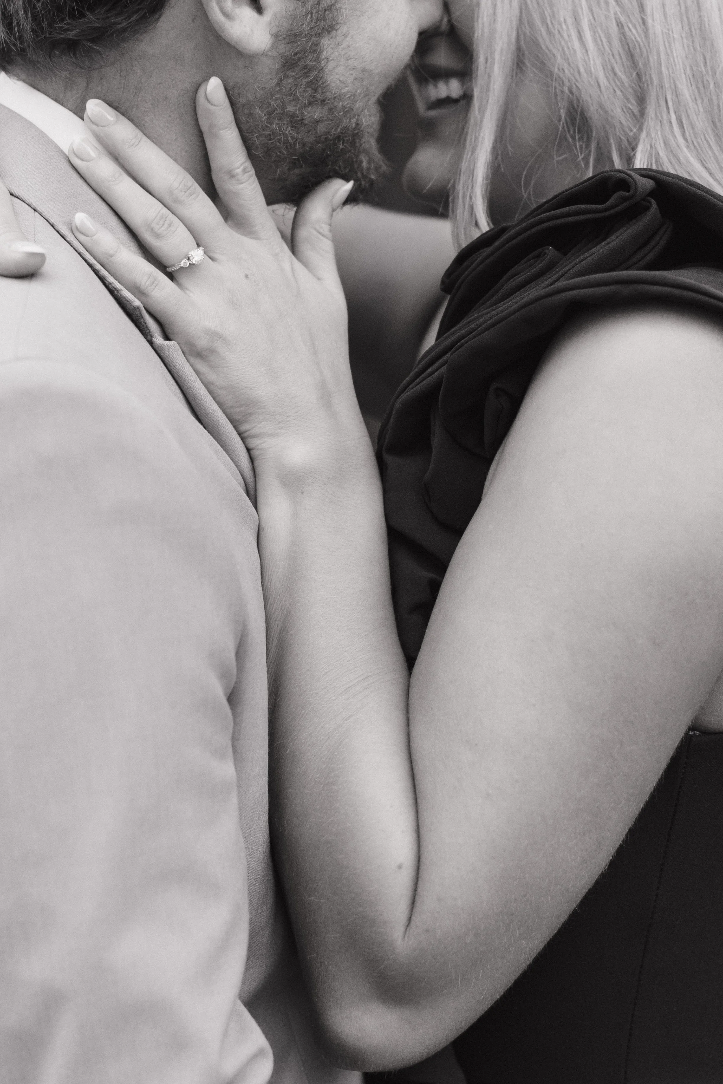 A close-up black-and-white photo of a couple touching faces and smiling, with the woman's hand resting on the man's neck, showing an engagement ring.