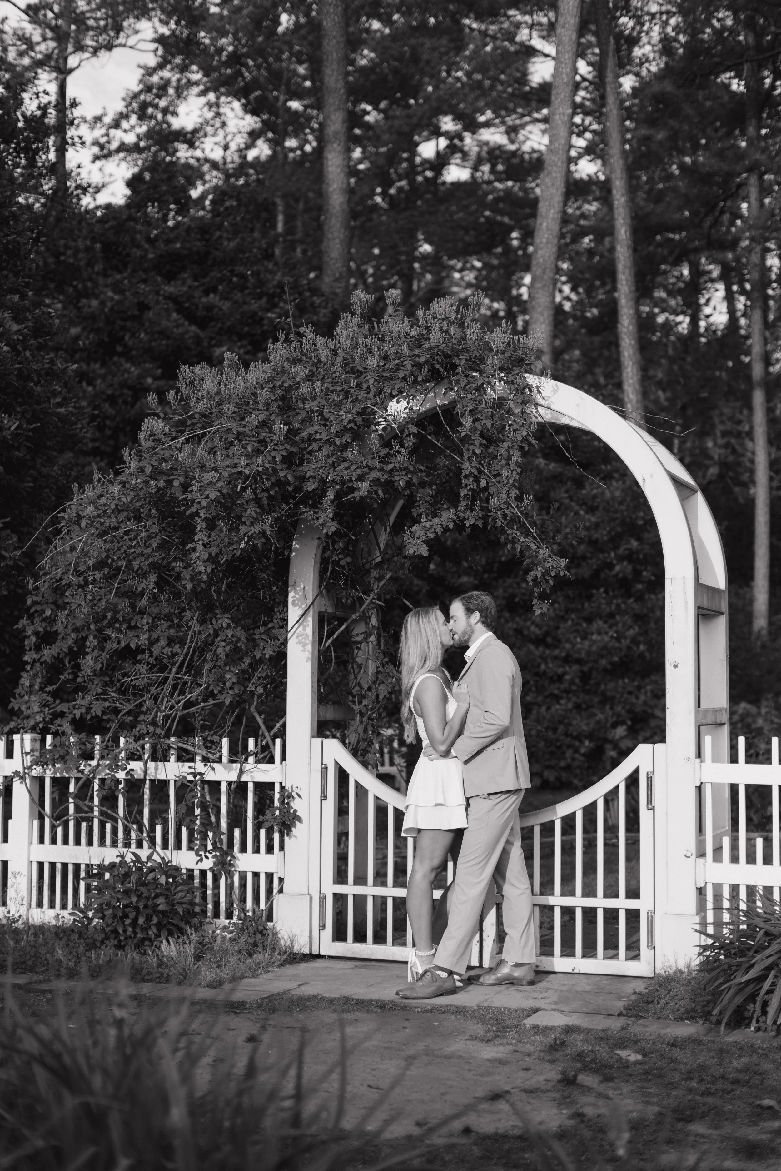 A couple dressed in formal attire standing close together, about to kiss, under a garden arch with foliage, surrounded by trees and a white picket fence in the background, in a black and white photograph.