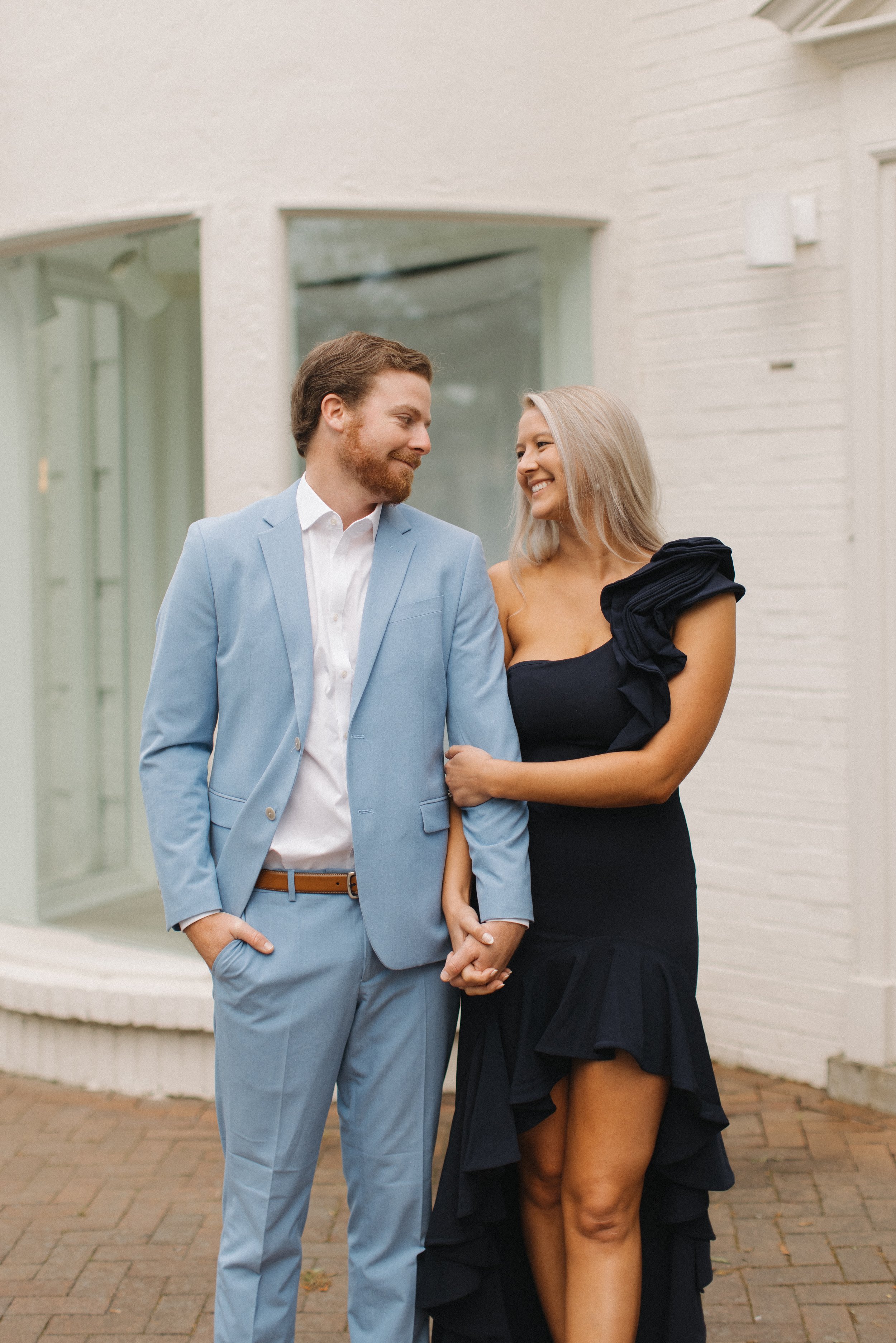 A man and woman standing outside, holding hands, smiling at each other.