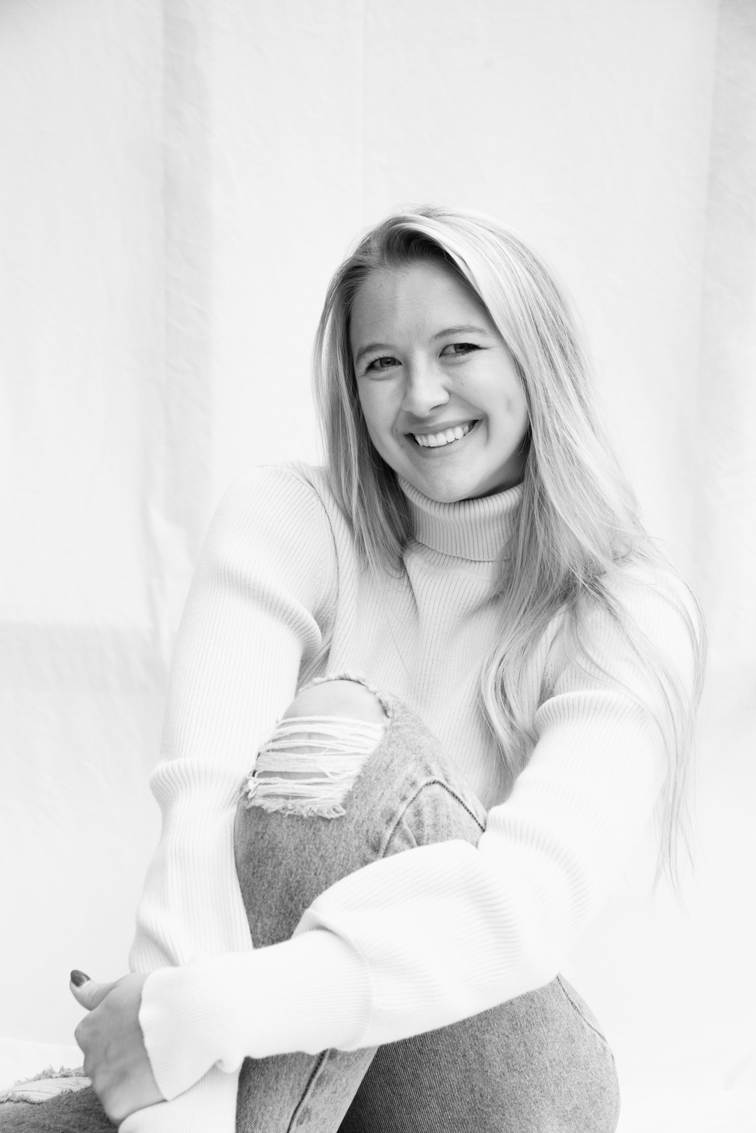 Black and white photo of a smiling young woman with long hair, wearing a turtleneck sweater and ripped jeans, sitting with her knee up and arm resting on it.