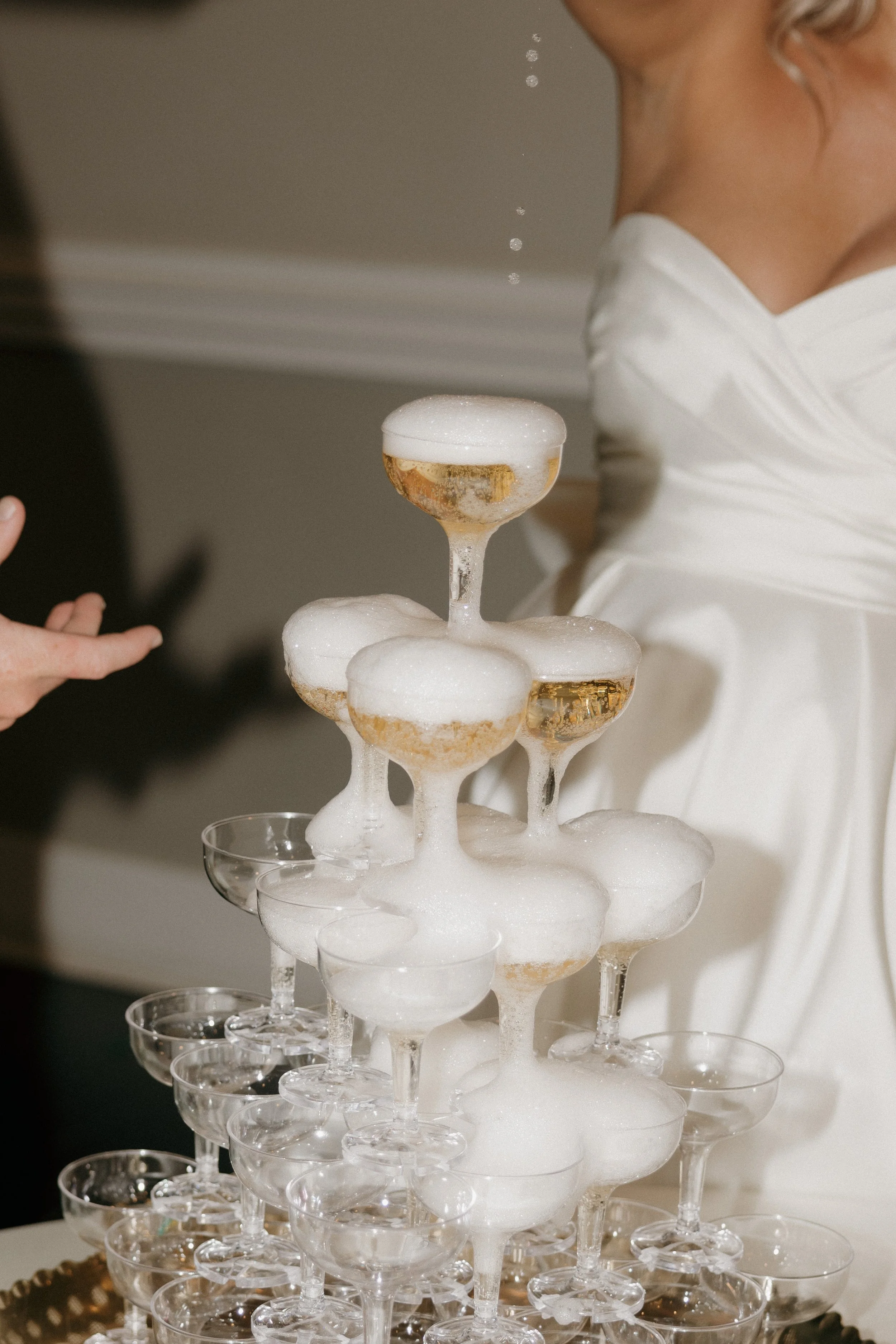 A champagne tower with glasses filled with bubbly, topped with foam, and a person dressed in white near it.
