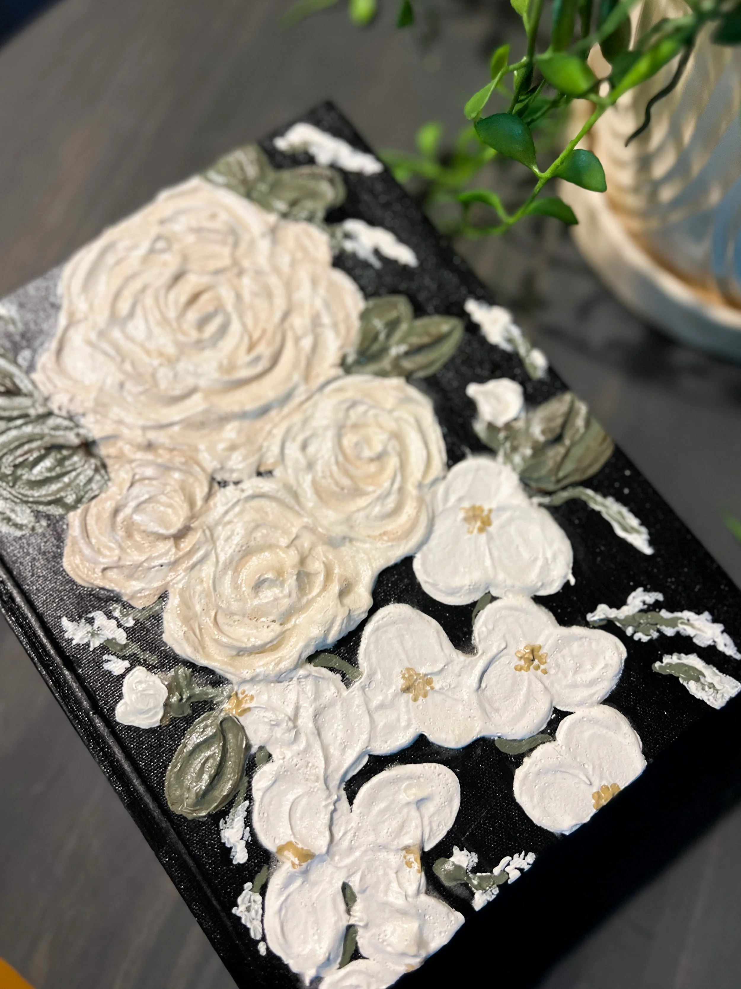 Decorative art piece with white and beige 3D flowers and green leaves on a black background, placed on a table near a plant.