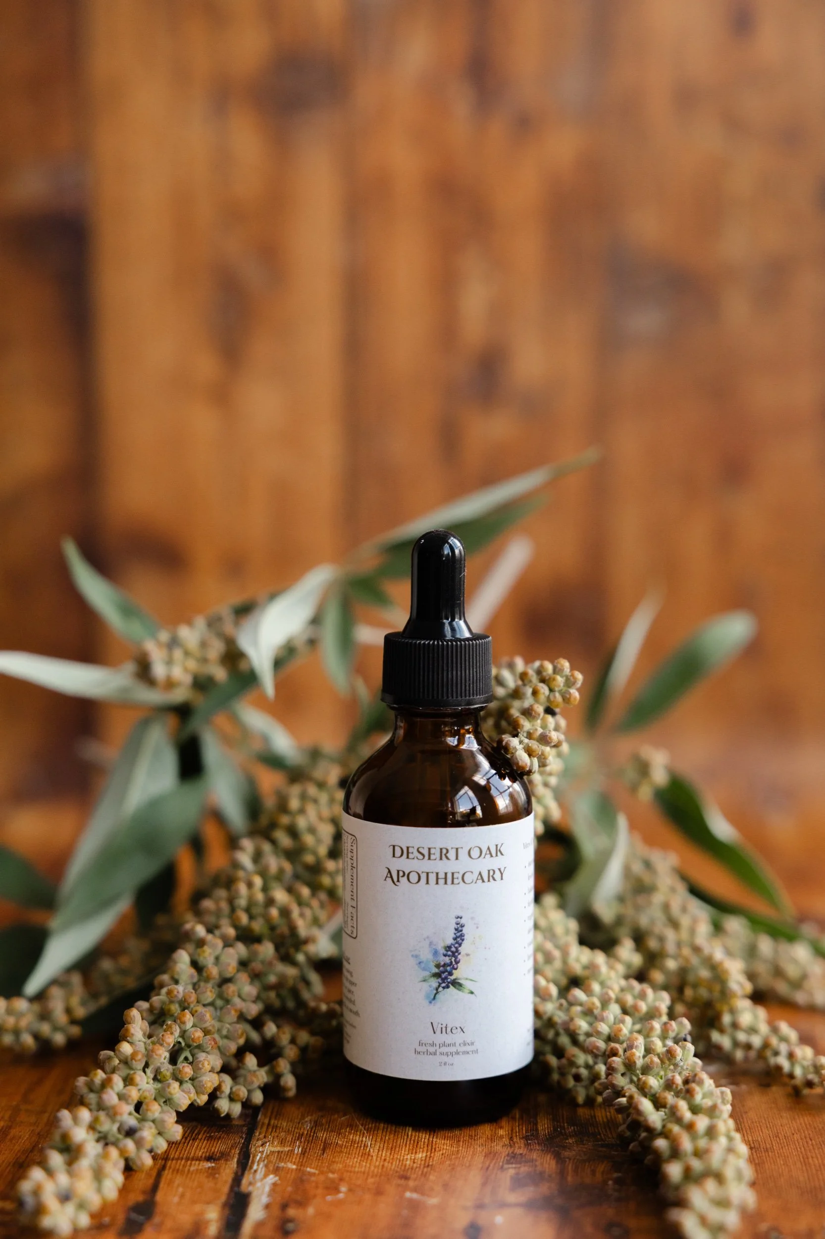 Amber glass bottle of Desert Oak Apothecary Vitex tincture with a black dropper, nestled among green leaves and dried flower clusters against a warm wooden background
