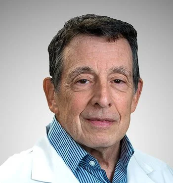 A middle-aged man with dark hair, wearing a white coat and a blue striped shirt, smiling lightly against a plain background.