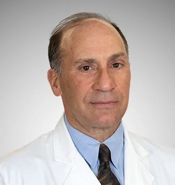 A middle-aged man in a white lab coat, light blue shirt, and patterned tie, looking directly at the camera against a plain background.