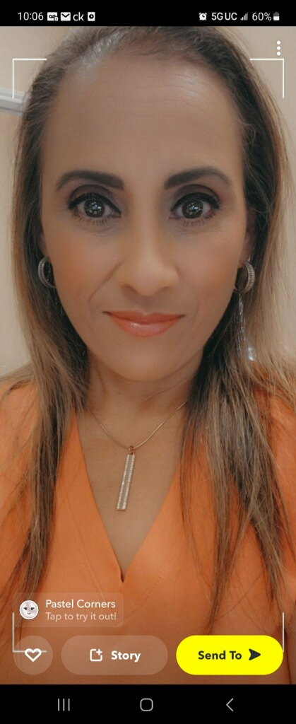 A woman with long, light brown hair, wearing a coral top, silver earrings, and a silver necklace with a vertical pendant. She has makeup on and is smiling at the camera.