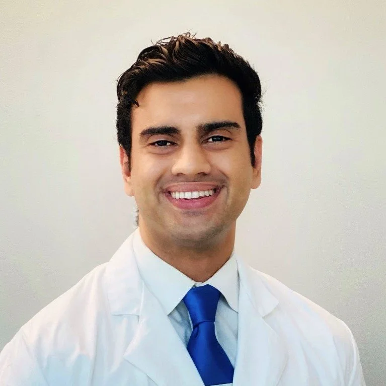 A smiling man in a white medical coat and blue tie against a plain light-colored background.
