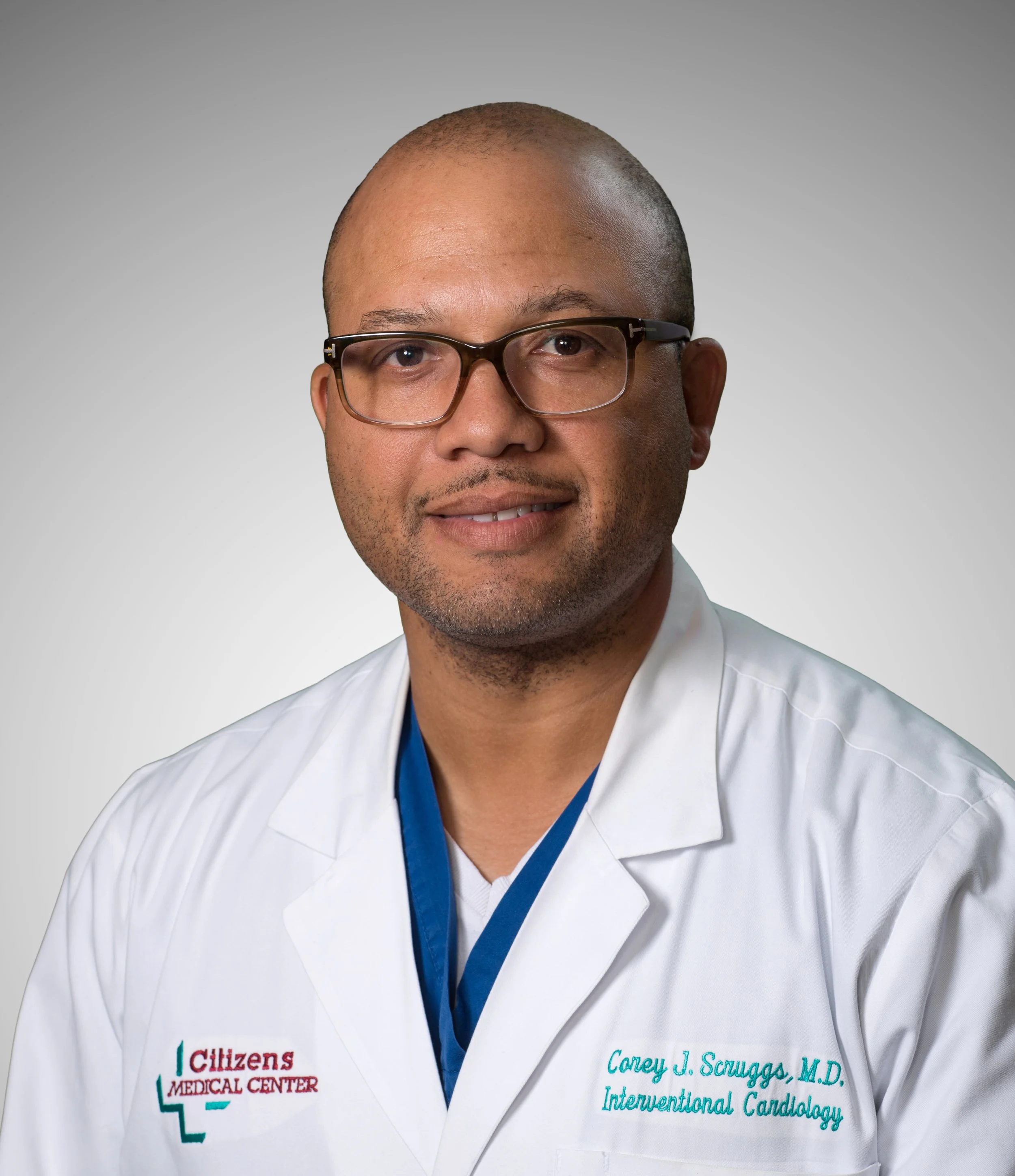 A man wearing glasses and a white lab coat with 'Citizens Medical Center' embroidered on it, smiling against a gray background.
