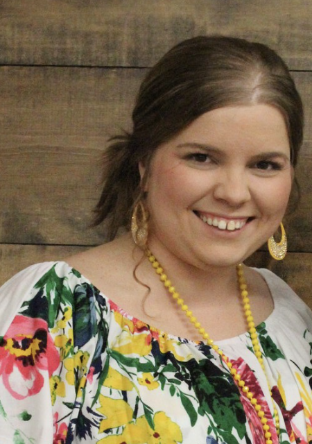 A young woman smiling, wearing large gold earrings, a yellow beaded necklace, and a white floral top with pink, yellow, and green colors, standing against a wooden background.