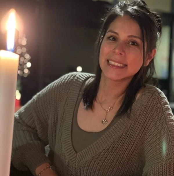 A woman with dark hair smiling at the camera, wearing a beige sweater and layered necklaces, sitting near a lit candle.