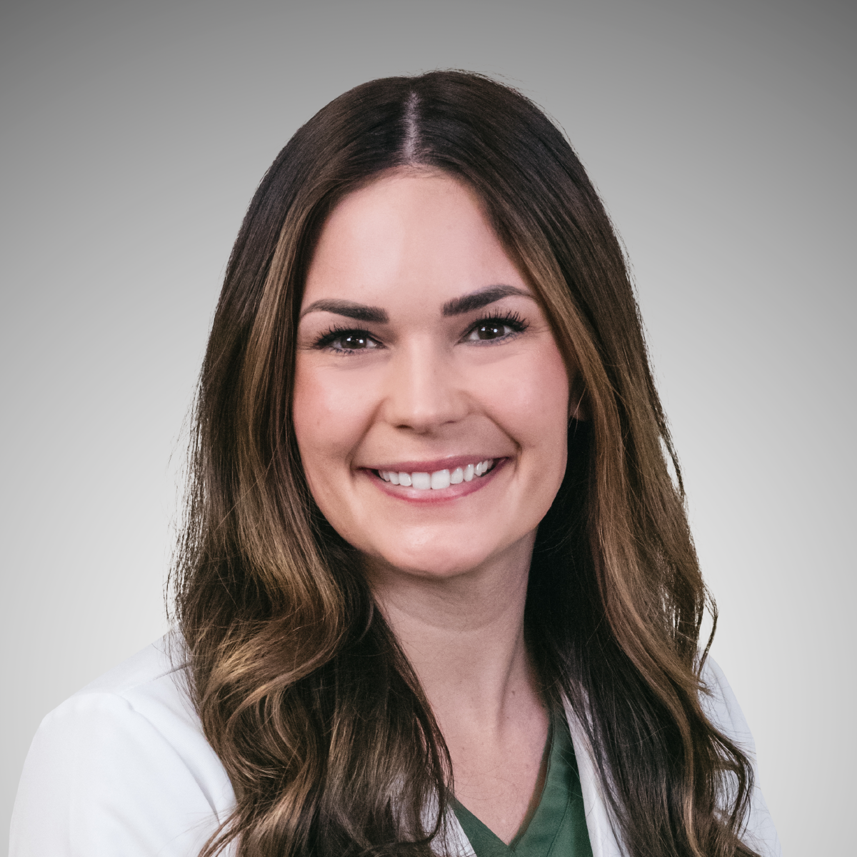 Portrait of a smiling woman with long wavy brown hair, wearing a white coat over green scrubs, against a neutral gray background.