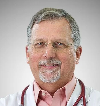 A middle-aged man with gray hair, glasses, and a beard, smiling, wearing a white lab coat and a pink shirt, with a stethoscope around his neck.