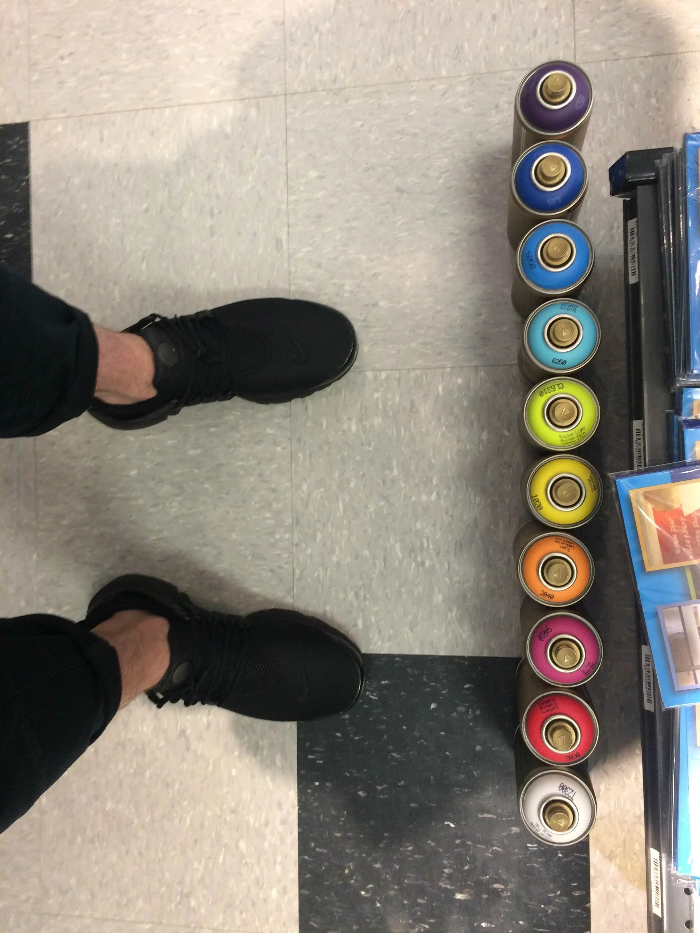 Line of rainbow colored spray paint cans on gray tiled floor in front Brian Madden's shoes
