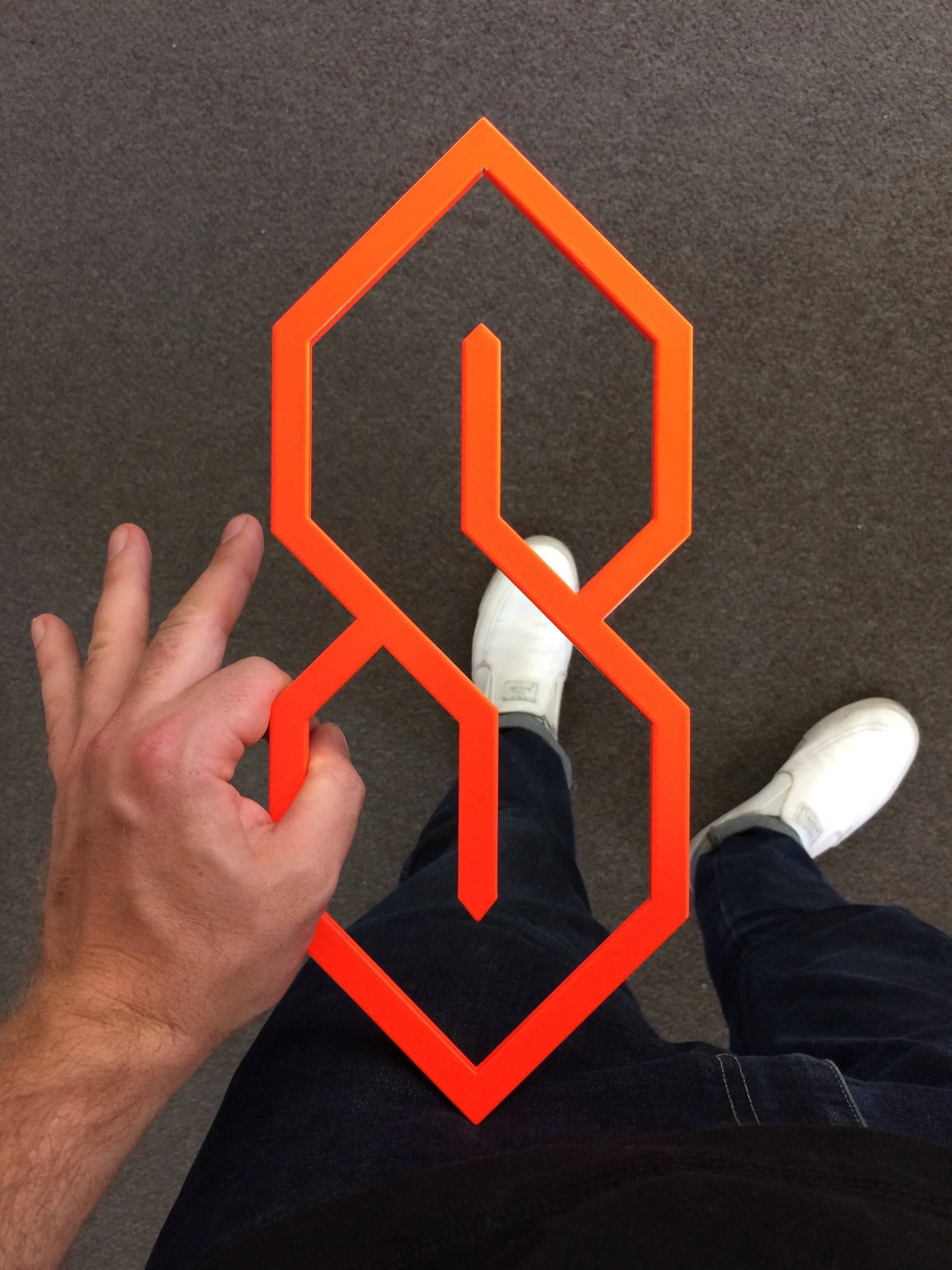 Artist Brian Madden's hand holding a flourescent orange powdercoated S-Thing trivet above a dark gray carpet floor