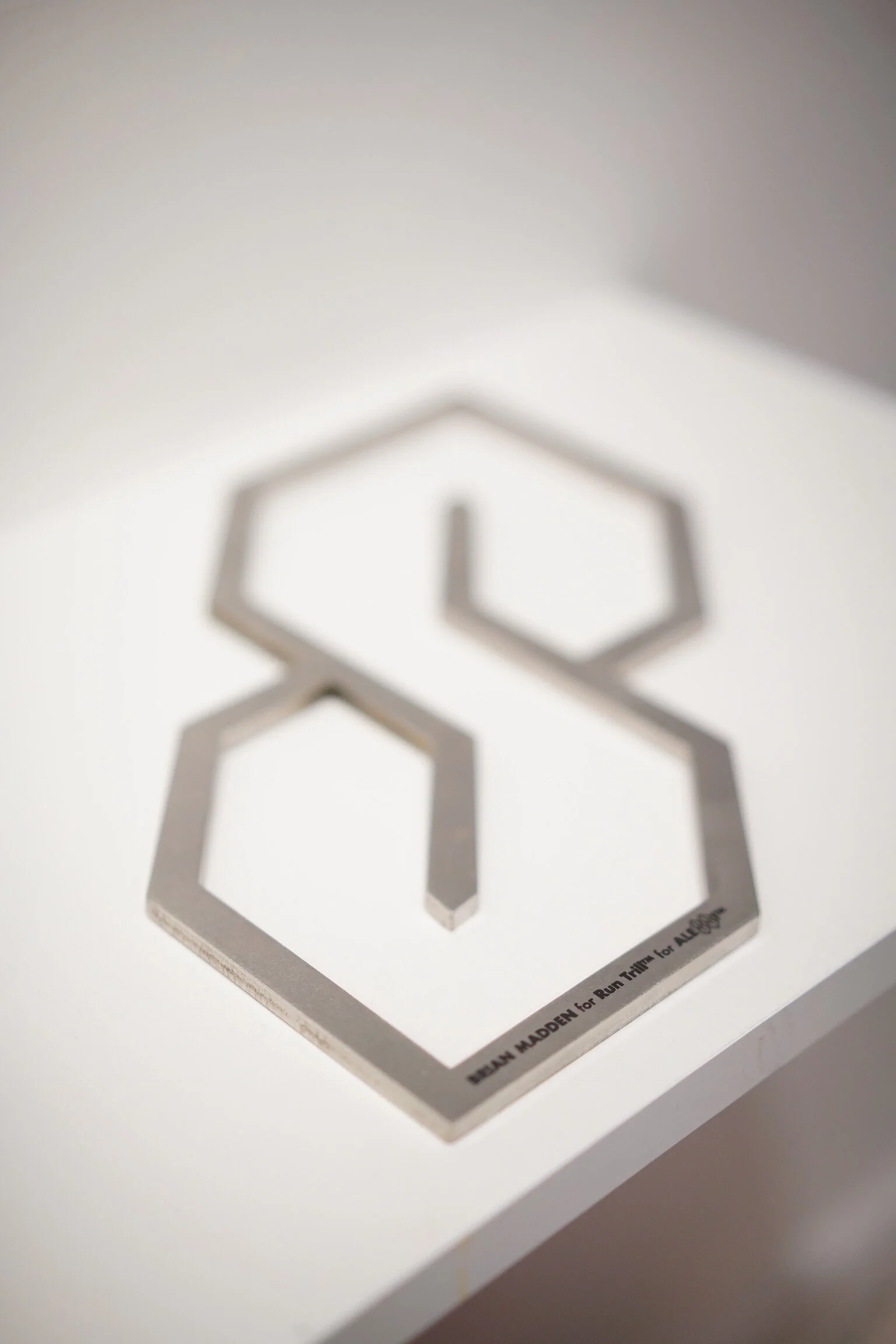 Stainless steel S-Thing trivet inspired by Alessi with black laser engraved Run Trill and Brian Madden branding sitting on a white wooden shelf