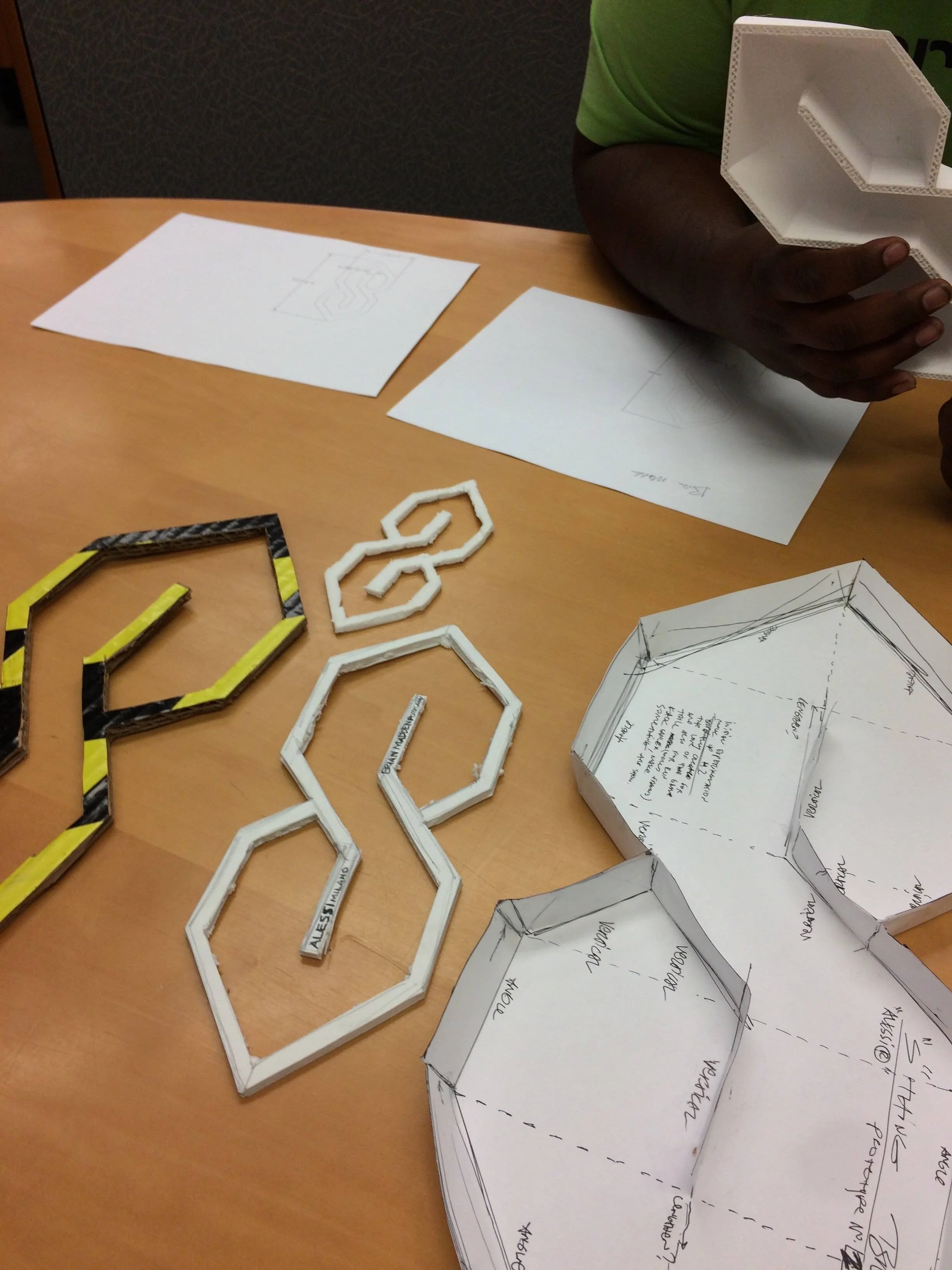 Three S-thing trivet and bowl designs made out of cardboard and foam on a light wood table in front of a black man's arm holding a white 3D-printed S-thing bowl