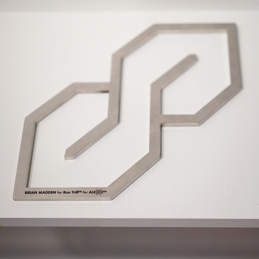 Stainless steel S-Thing trivet inspired by Alessi with black branding sitting on a white wooden shelf