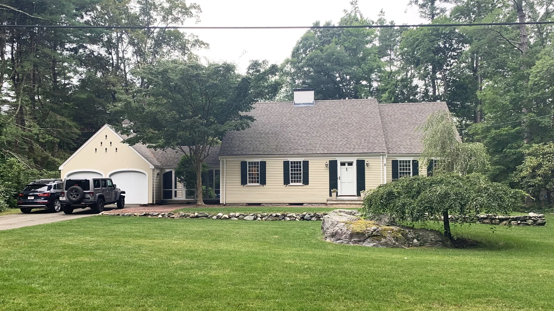Suburban house with attached garage, two parked SUVs, well-maintained lawn, trees, and stone landscaping.