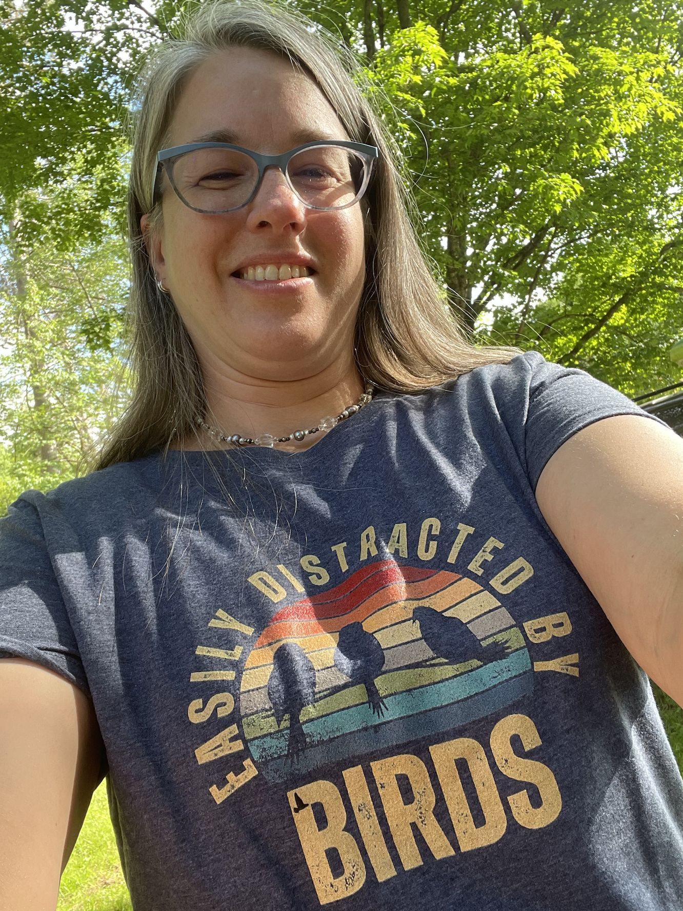 A woman outdoors in a green, leafy area taking a selfie. She is wearing glasses, a necklace, and a navy blue T-shirt with a bird graphic and the words "Easily Distracted By Birds."