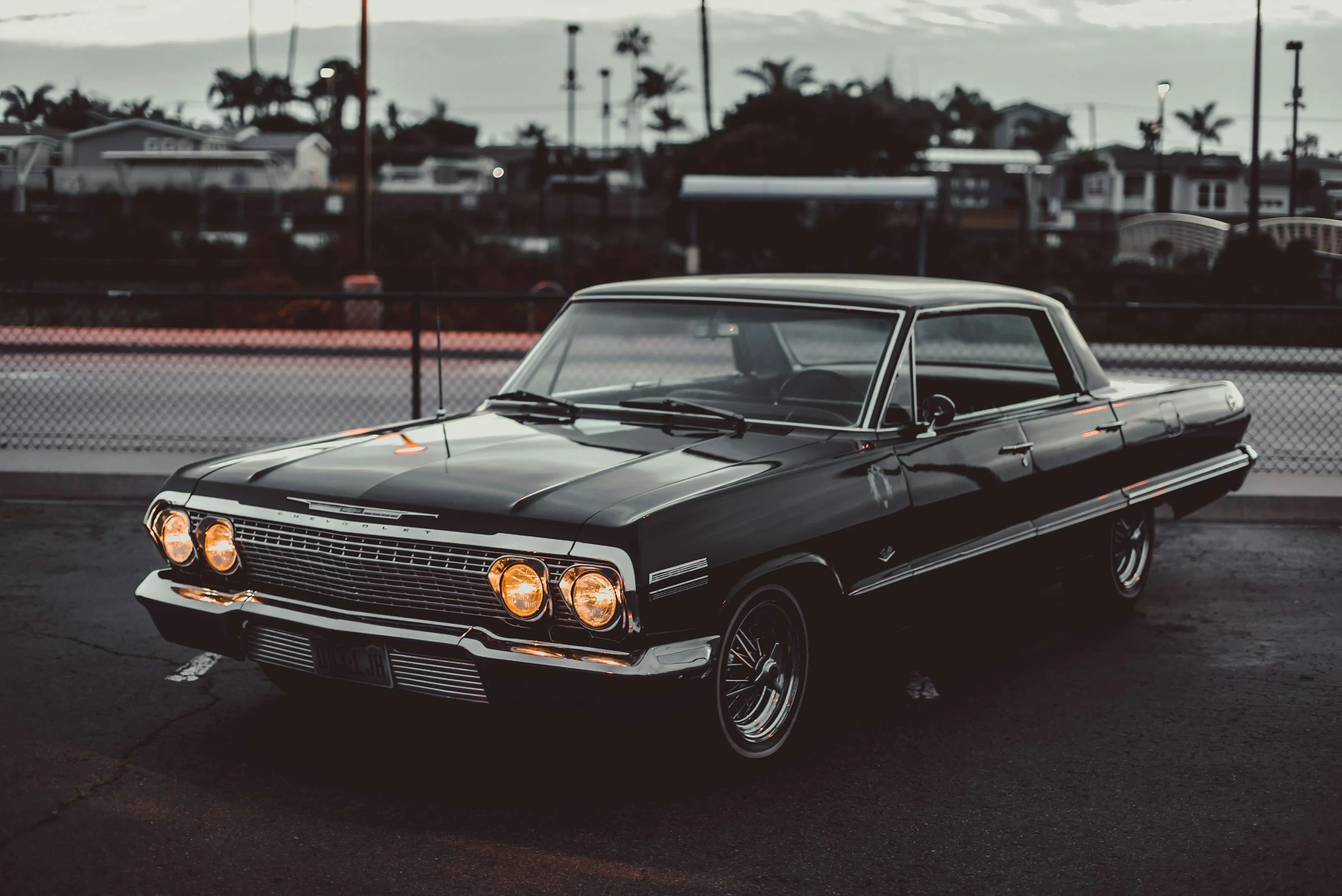 A vintage black car, likely from the 1960s, parked in a lot at dusk with some streetlights on. The background features a fence, residential houses, and palm trees.