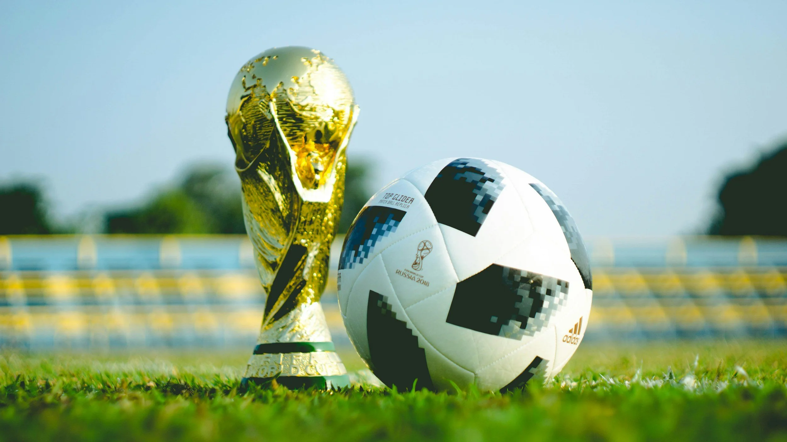 A soccer ball and a FIFA World Cup trophy placed on a grass field, with a blurred stadium in the background.