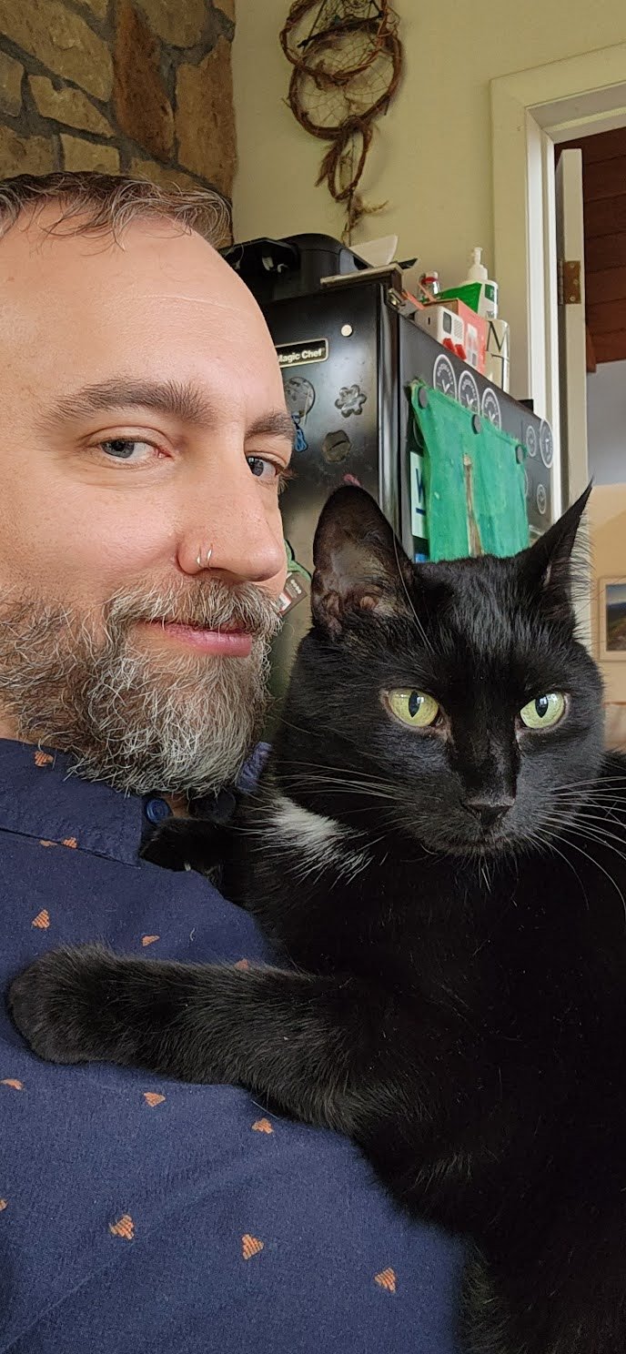 A man with a beard and a nose ring holding a black cat with green eyes indoors.