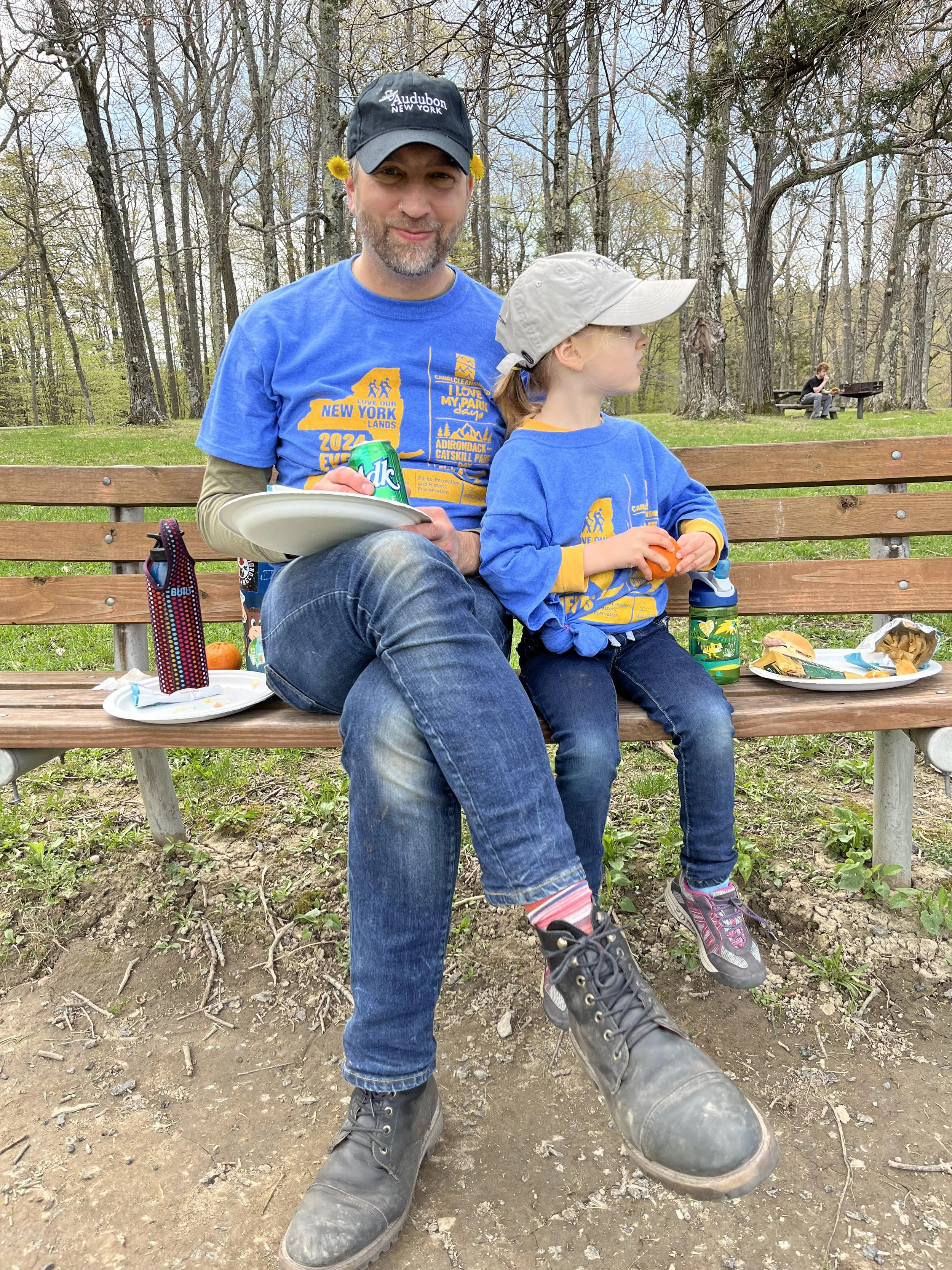 A man and a young girl sitting on a park bench with trees in the background. The man has a beard and is wearing a blue t-shirt, jeans, black boots, and a black cap with yellow flowers tucked behind his ears. They are having a picnic with food and drinks on the bench.