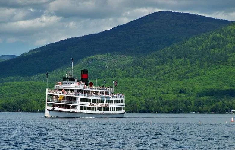 A large white steam-powered paddleboat with three levels floats on Lake George, with the Adirondacks in the background.