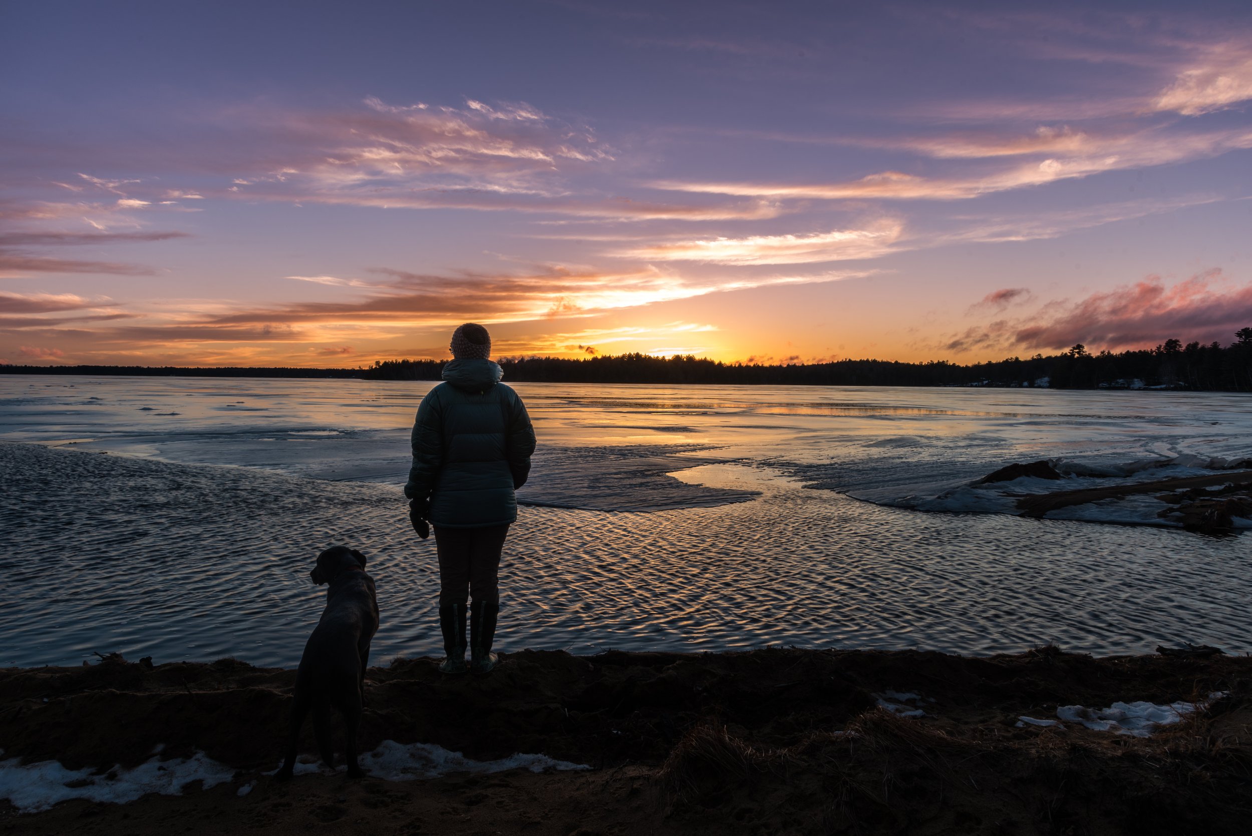 A person and their dog stand on a frozen Adirondack lakeshore, watching the sunset over the ice.